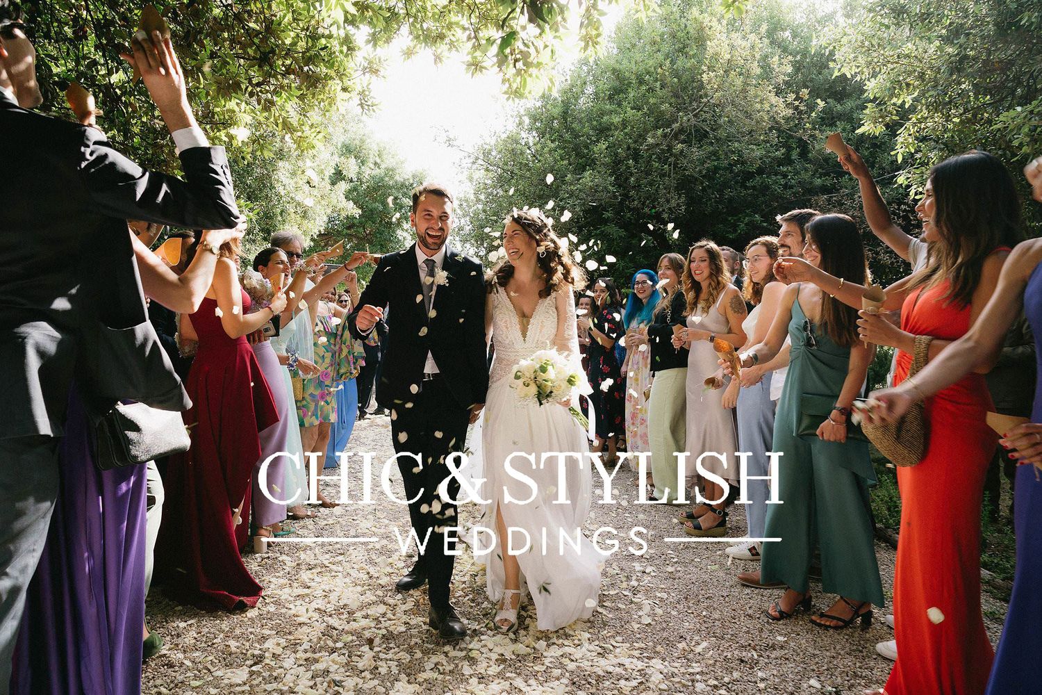 Wedding guests dance and celebrate together under trees at an outdoor garden reception during golden hour.