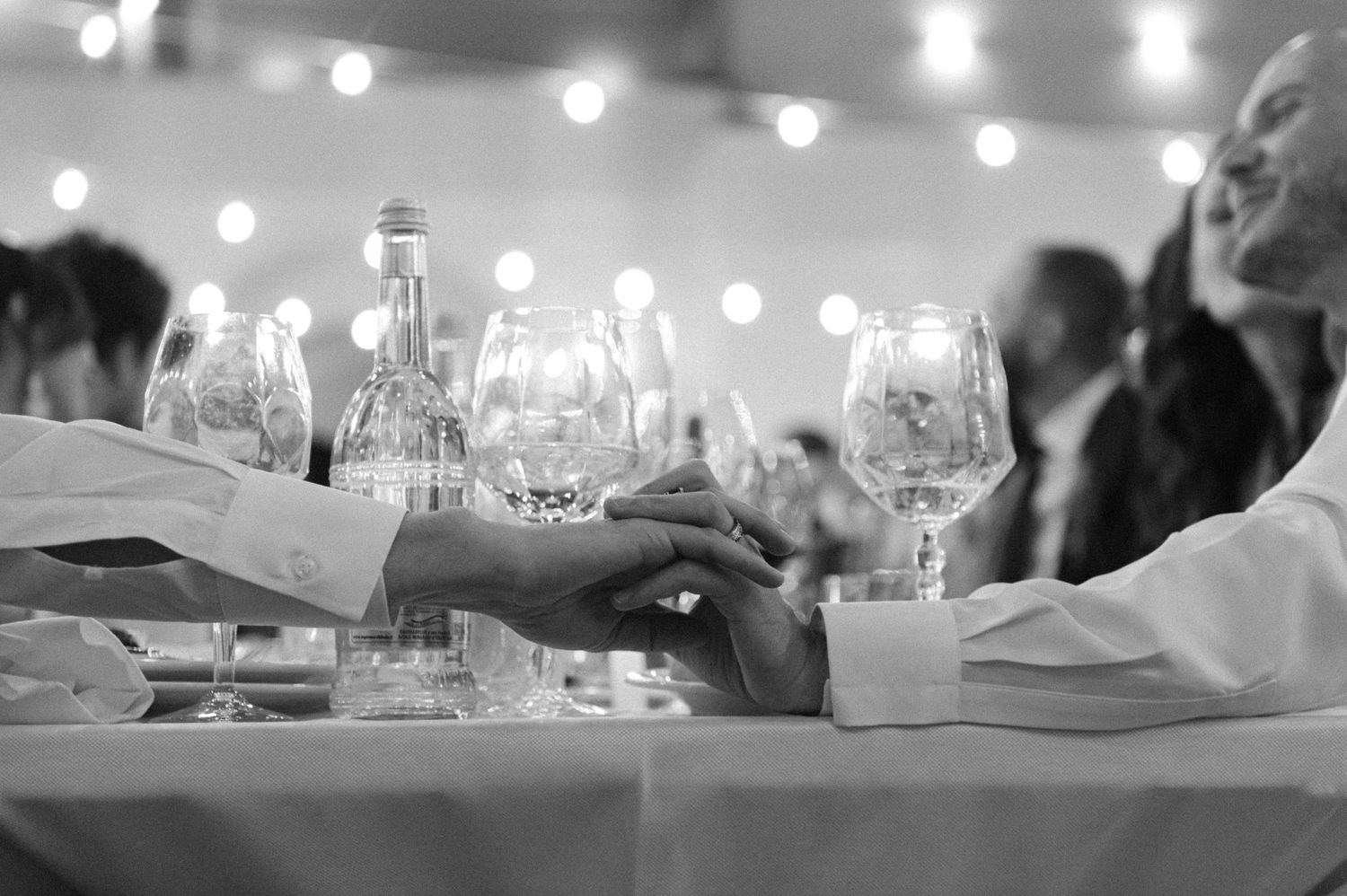 A black and white image of wine glasses on a dining table with string lights glowing softly in the background.