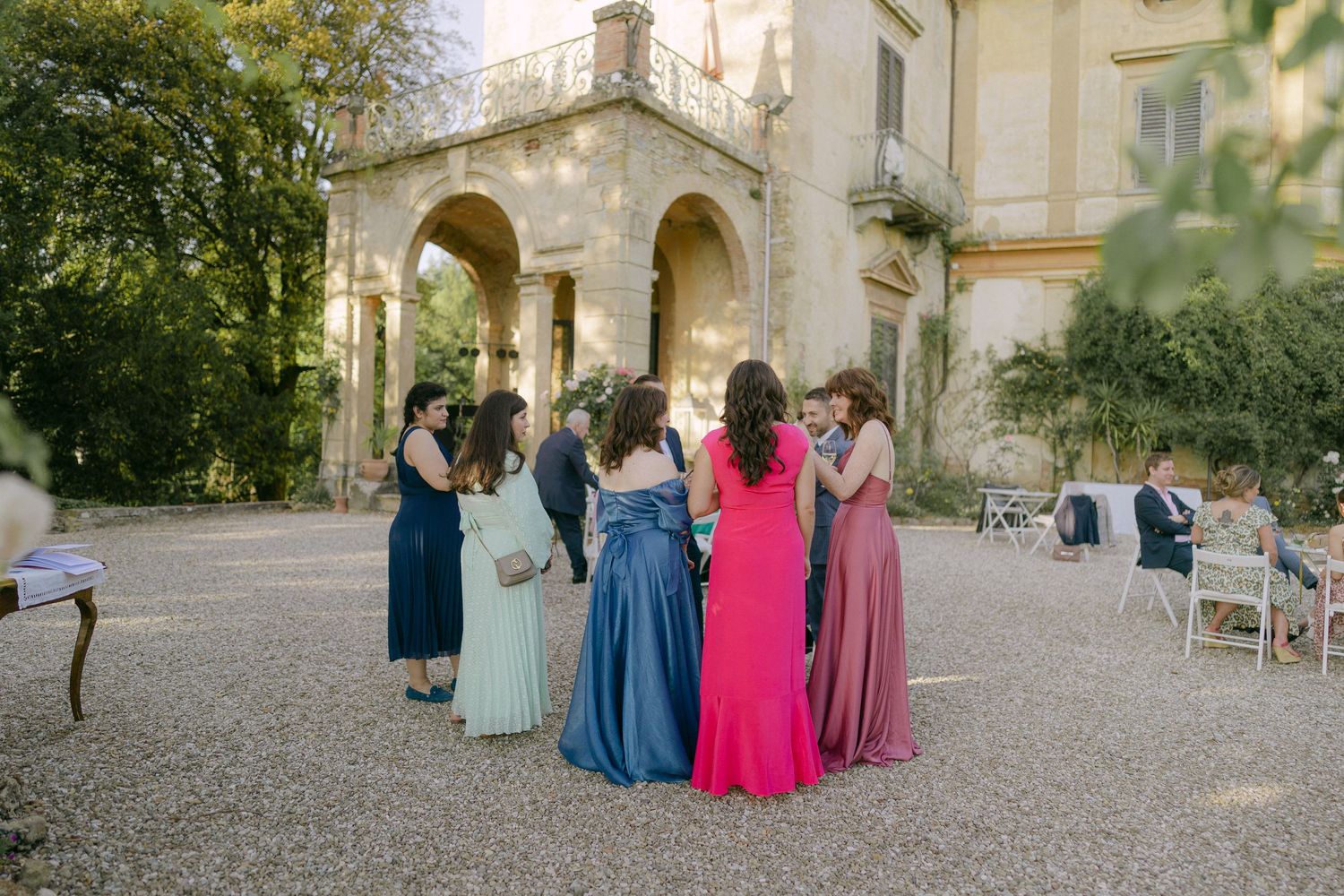 Wedding guests in colorful formal attire gather outdoors at an elegant stone venue for photos.