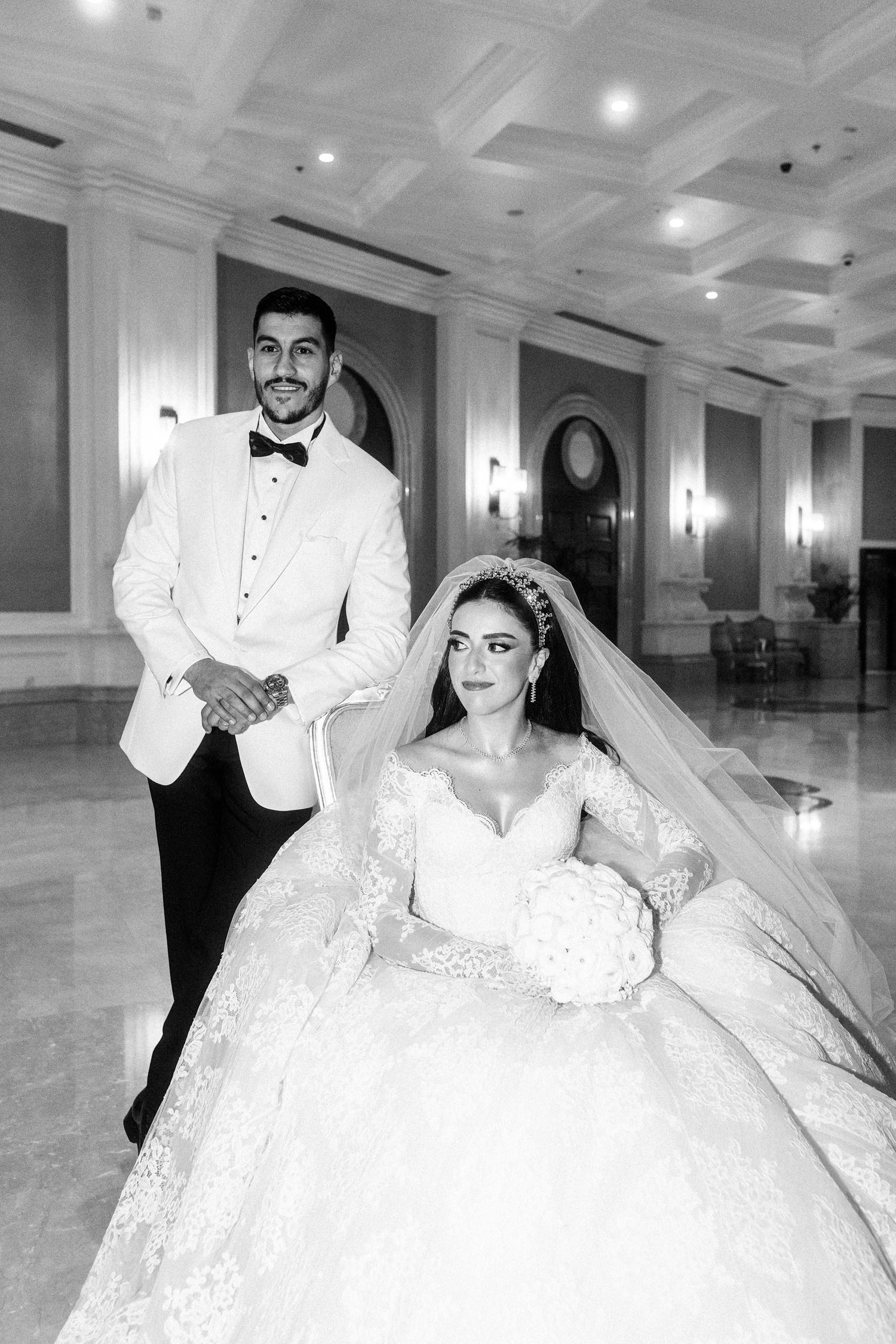 Black and white photo of a bride and groom at their wedding, she in an ornate lace gown and veil, he in a white tuxedo jacket.