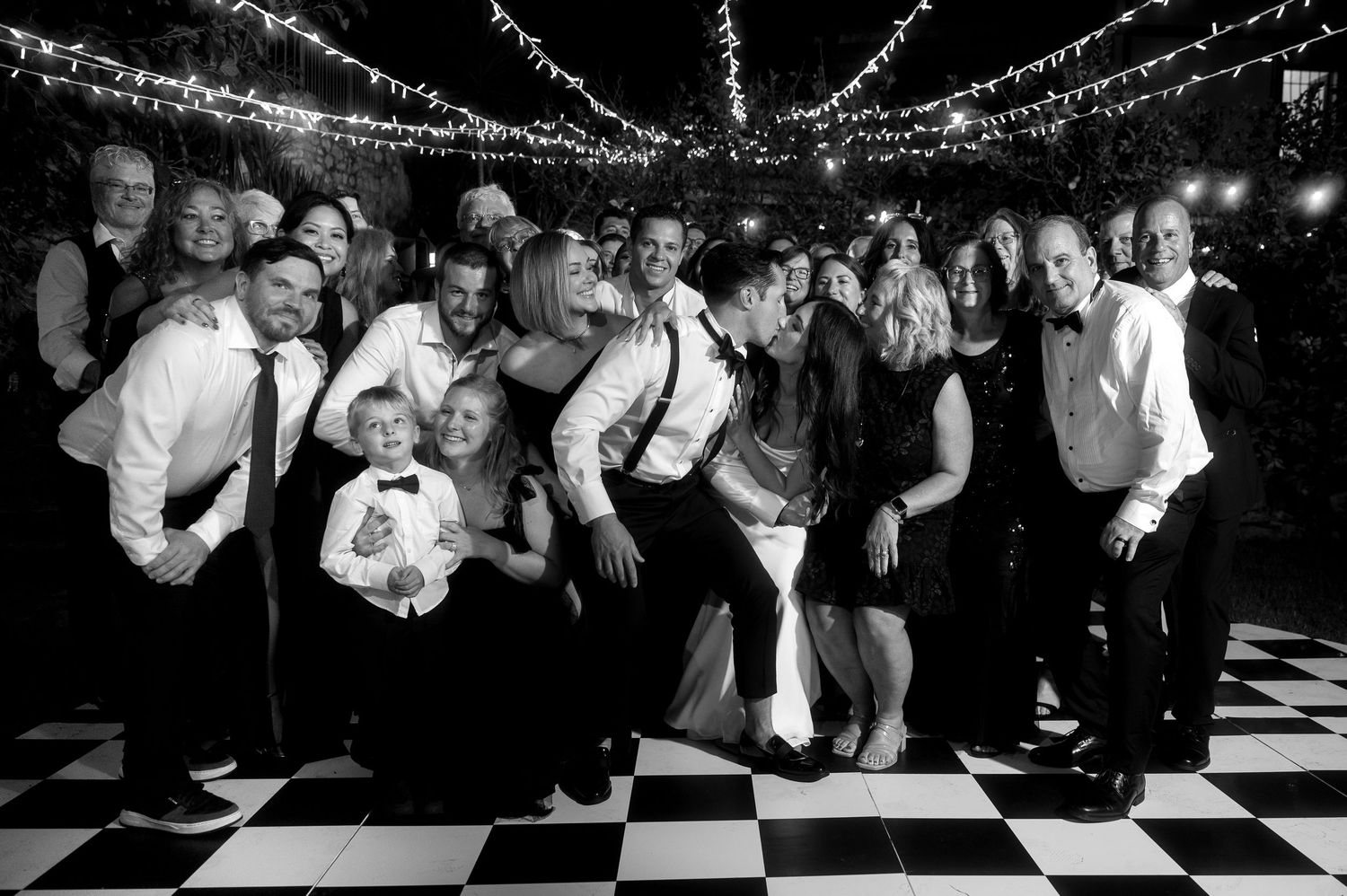Wedding guests dance together on a black and white checkered dance floor under twinkling lights at a reception celebration.