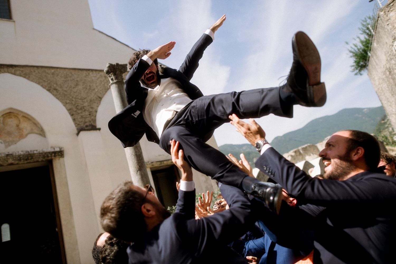 Wedding guests playfully tossing a groom in the air during outdoor celebration.