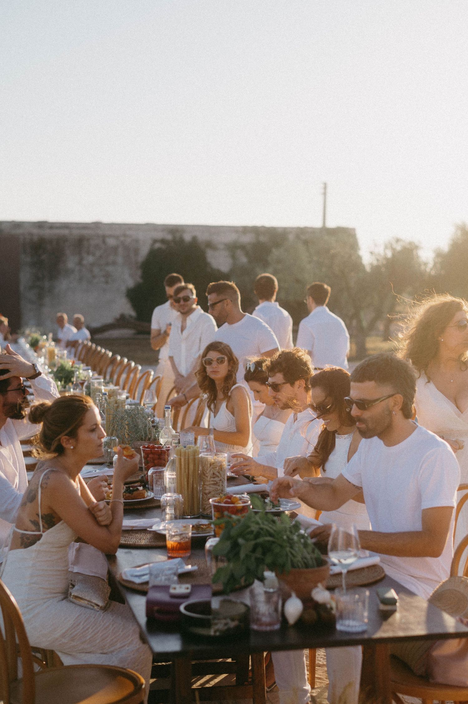 Sunset dinner party guests dressed in white enjoying a long outdoor table setting with glowing golden light.