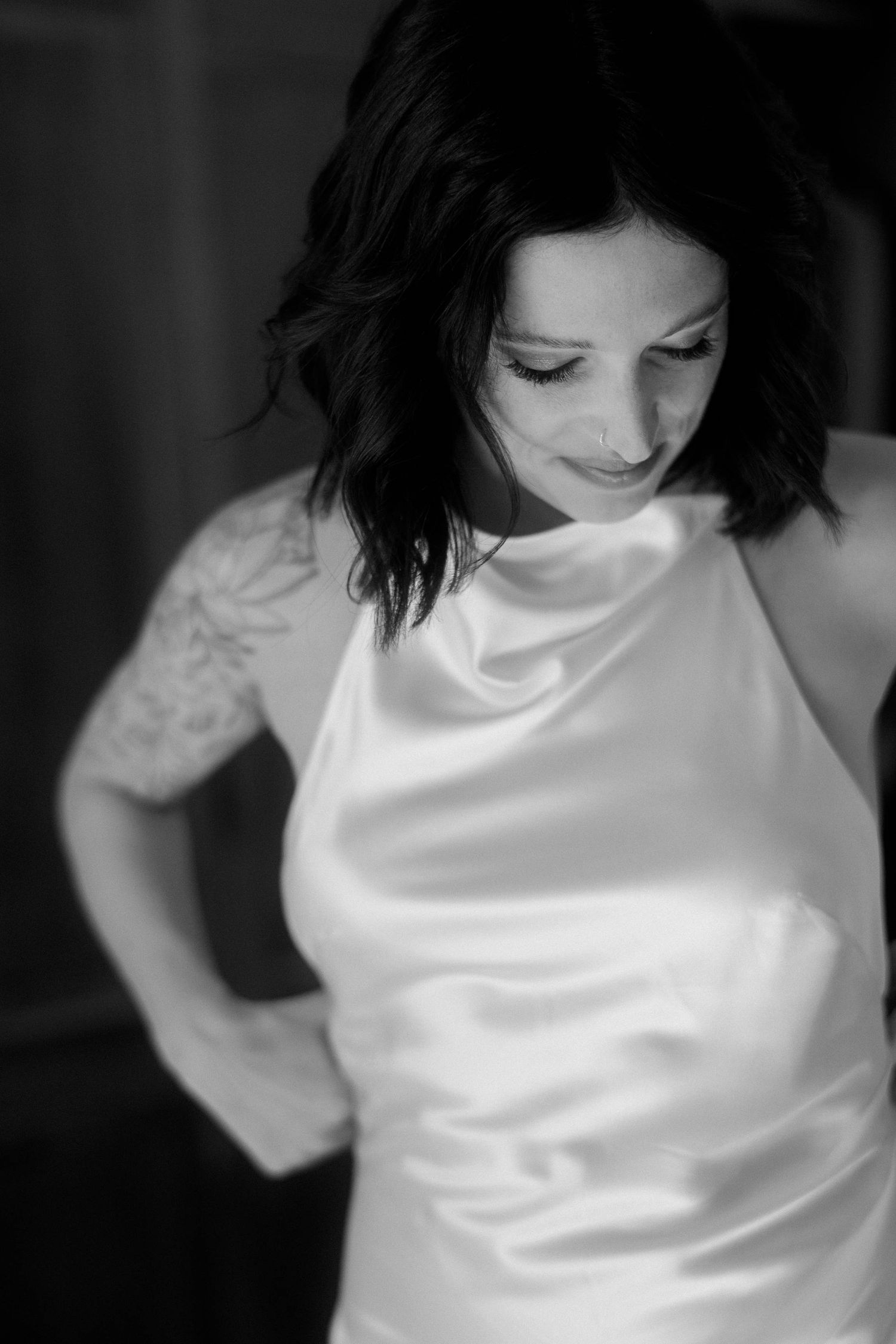 A black and white portrait of someone in a white satin top with lace sleeves looking down with a gentle smile.