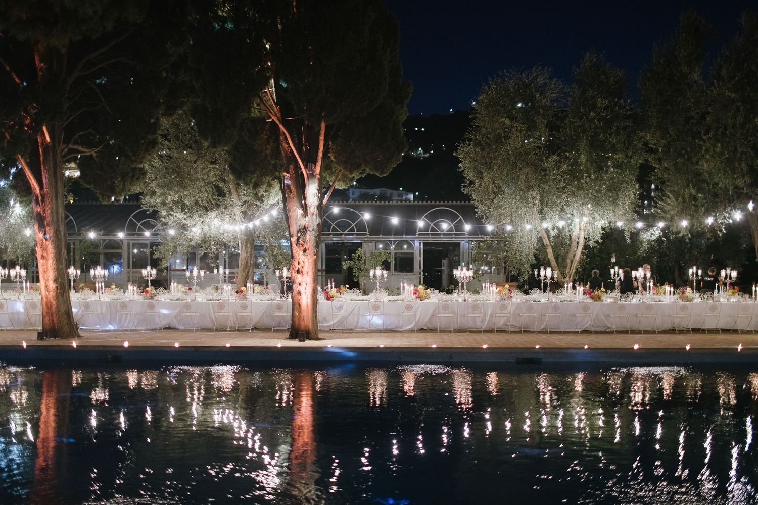 String lights illuminate a bridge and reflect off calm water at night creating a magical outdoor atmosphere.