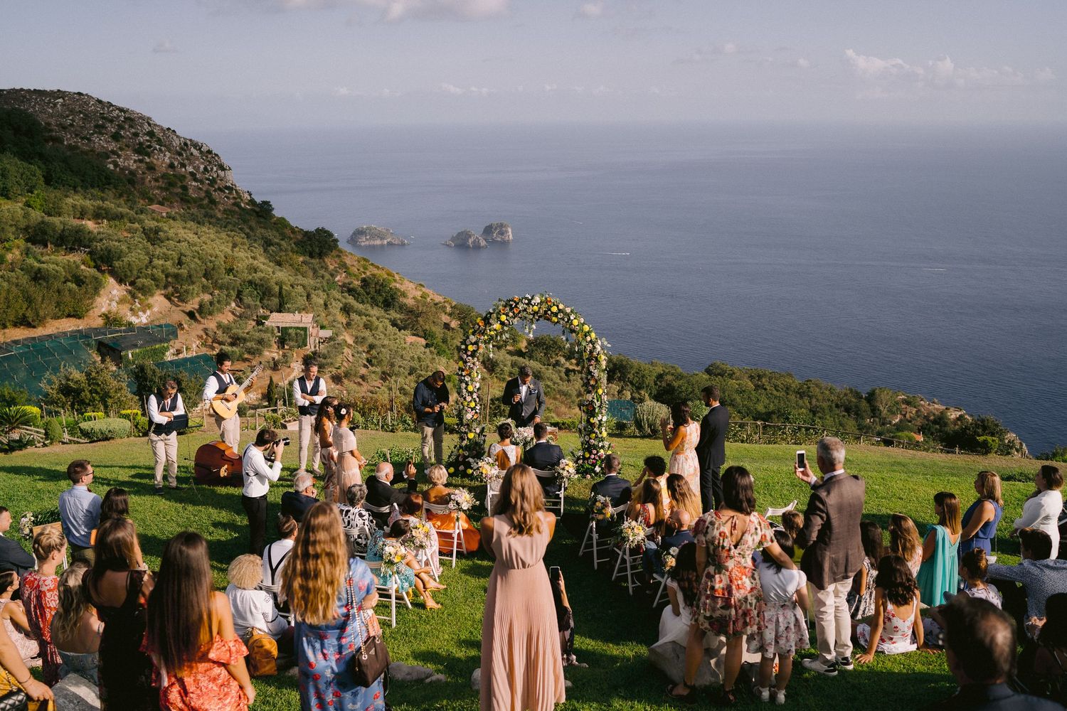 A scenic outdoor gathering overlooks the ocean from a hilltop with rock formations visible in the distance.