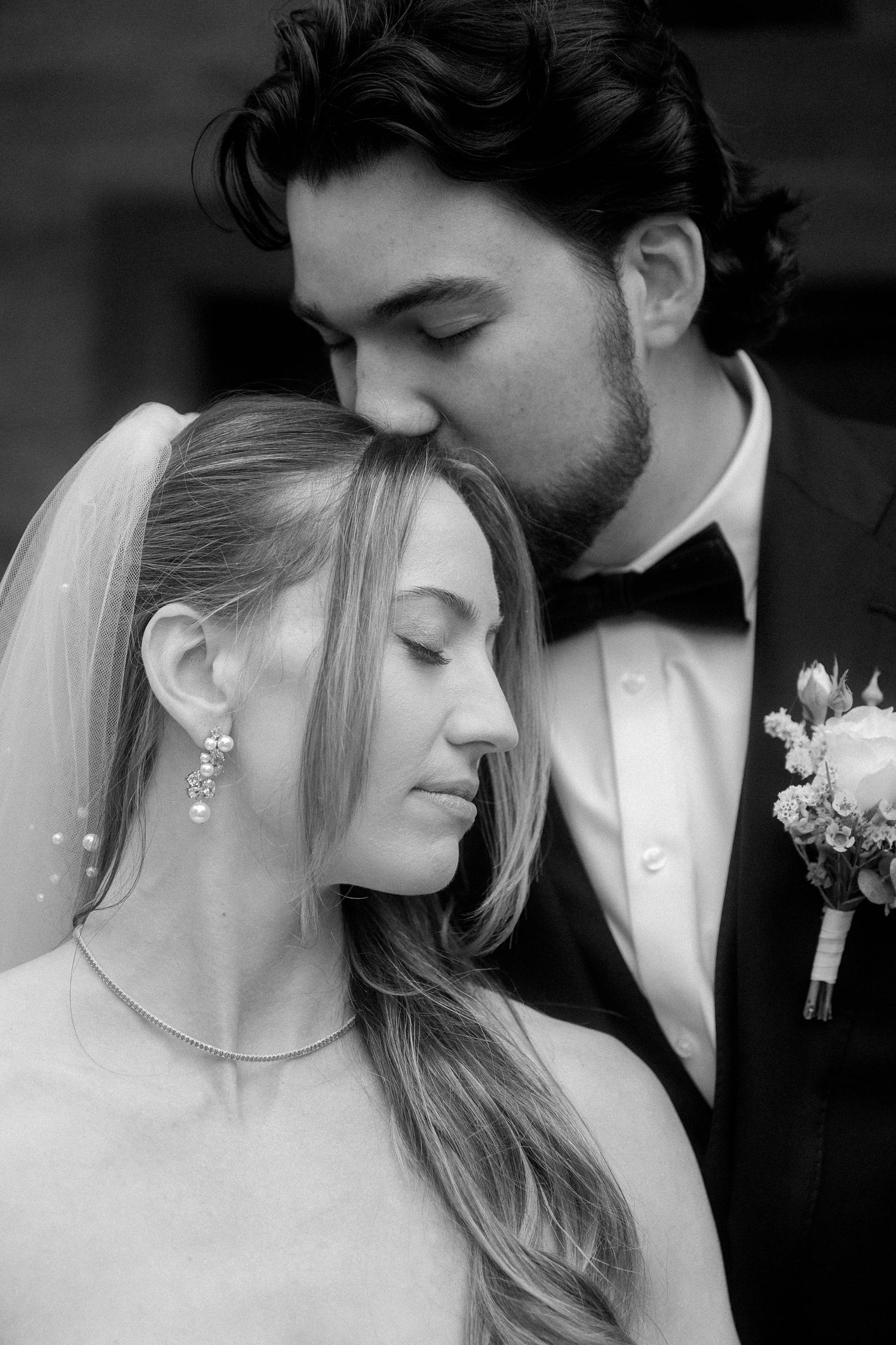Black and white wedding portrait of bride and groom, groom kissing bride's head, both in formal attire.