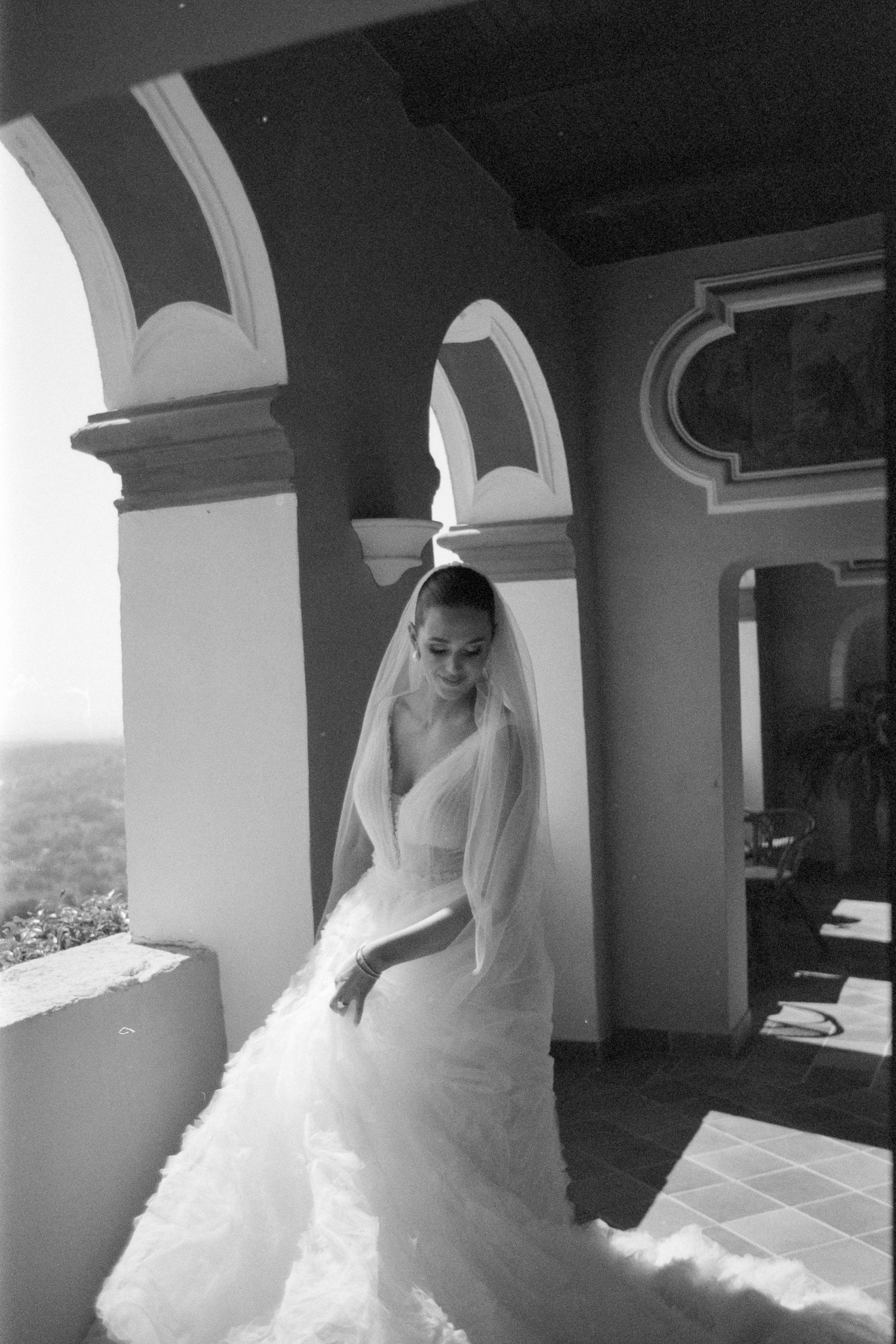 A bride in a white wedding gown poses elegantly in an arched corridor with scenic mountain views in the background.
