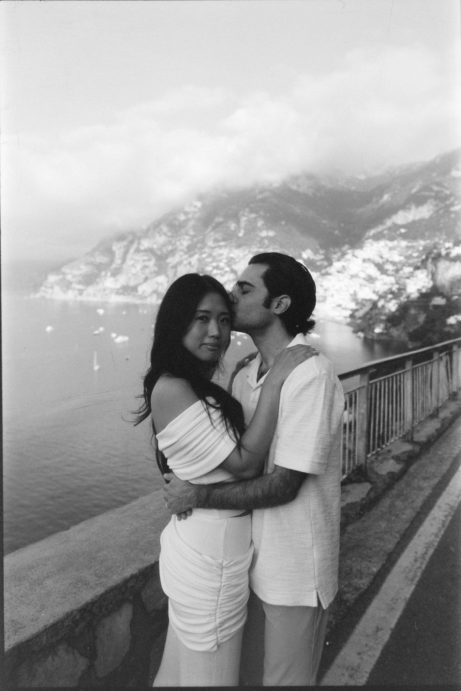 A romantic couple embraces while standing on an overlook with dramatic coastal mountains visible in the background.
