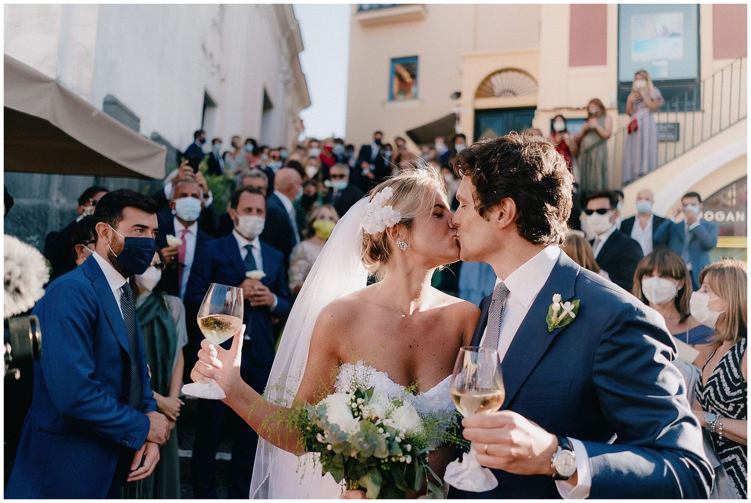 A bride and groom share a celebratory kiss while holding champagne glasses surrounded by guests at an outdoor wedding.