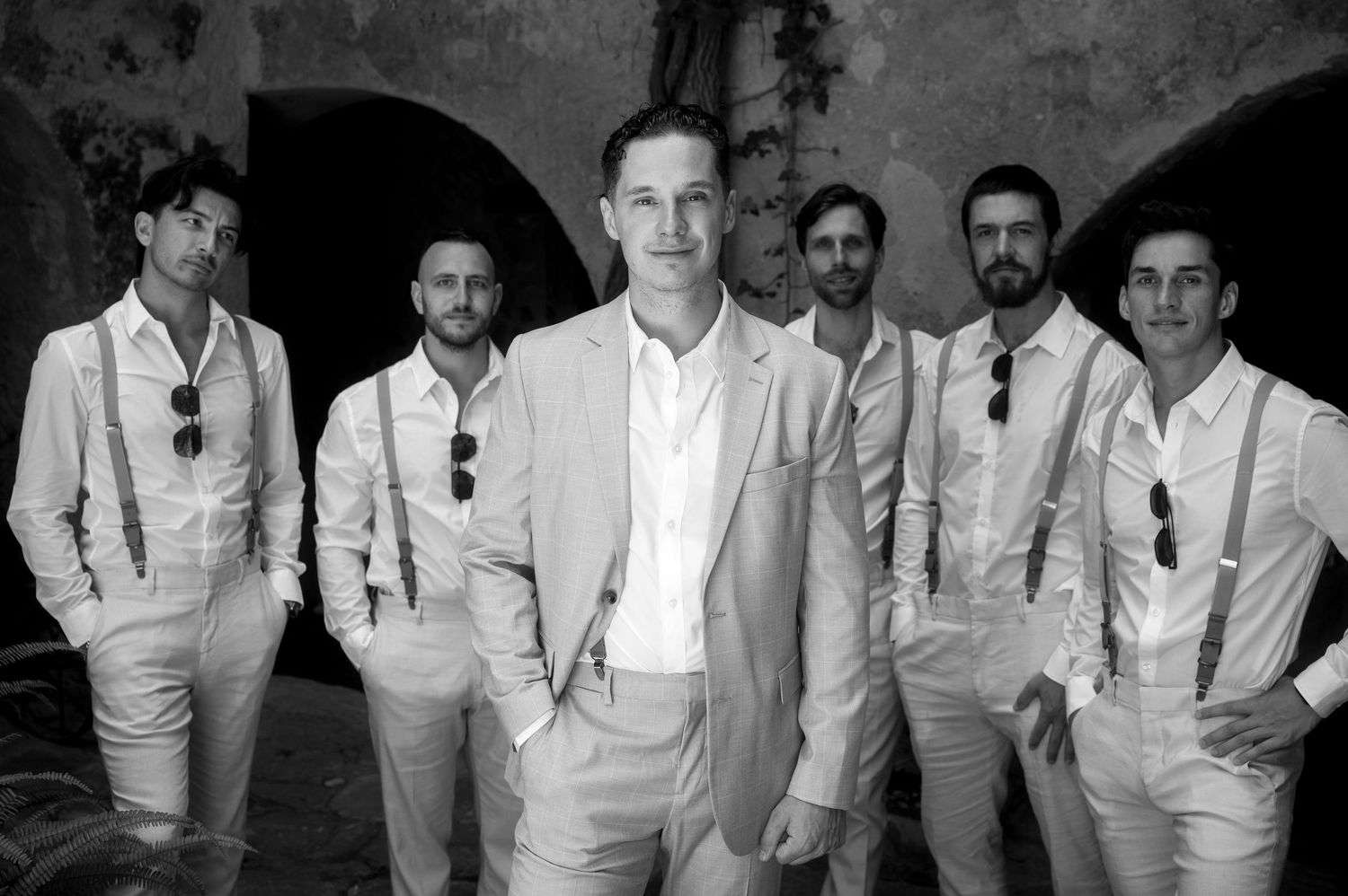 Black and white photo of a wedding party wearing light colored suits posing together against a stone wall.
