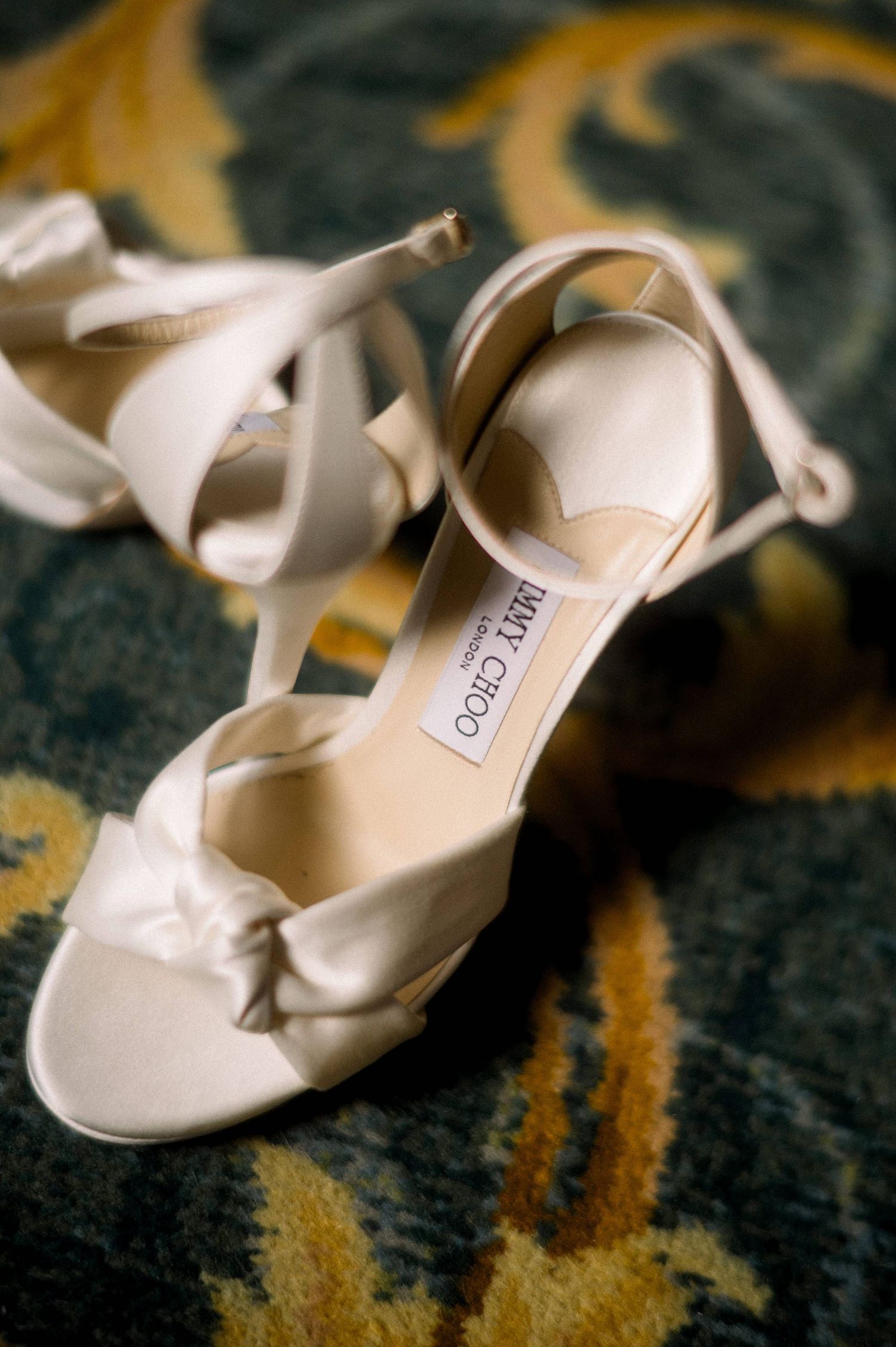 Elegant white Jimmy Choo bridal sandals with knotted straps resting on a patterned carpet.