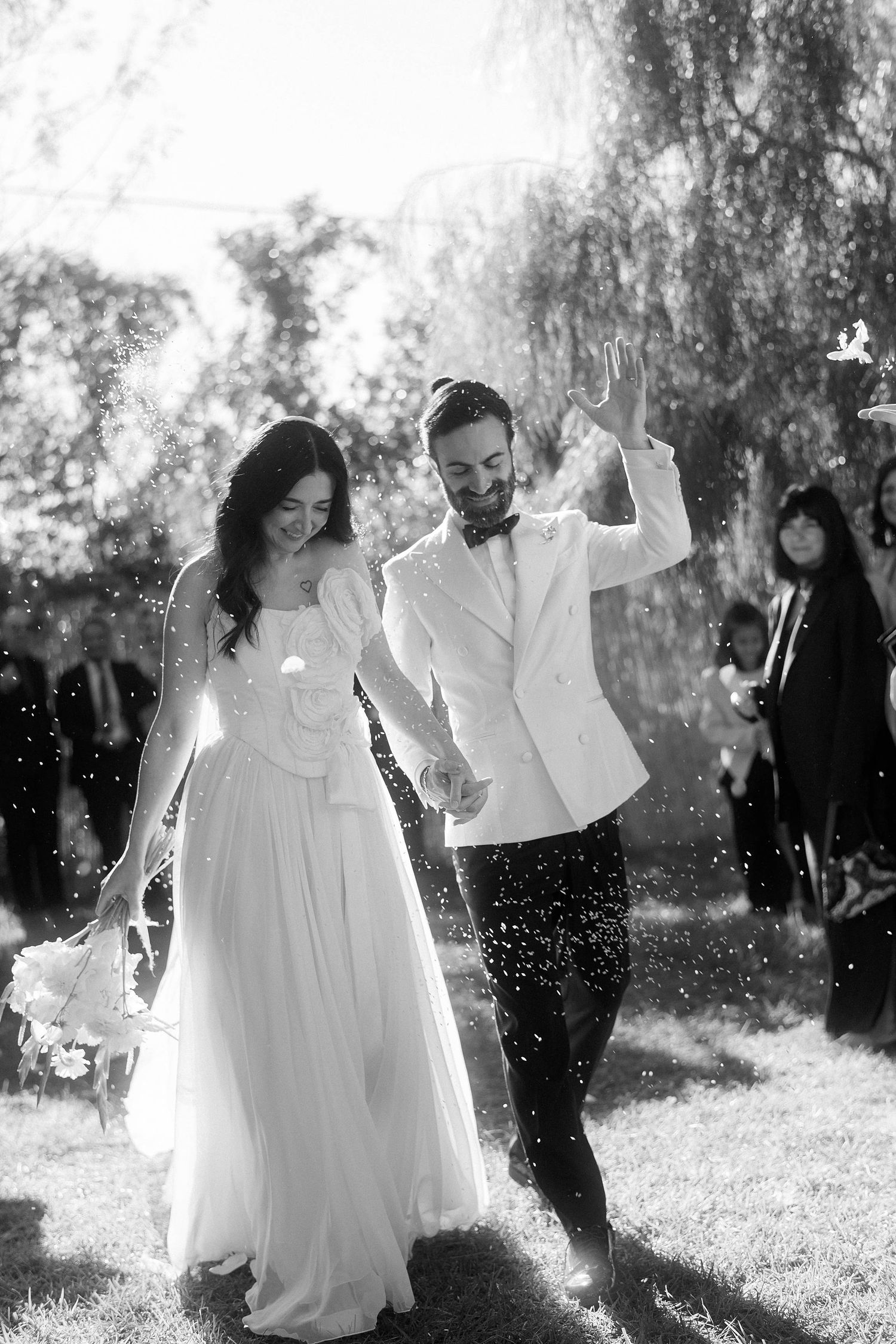 A joyful couple in wedding attire dance together outdoors on a sunny day with water droplets sparkling around them.