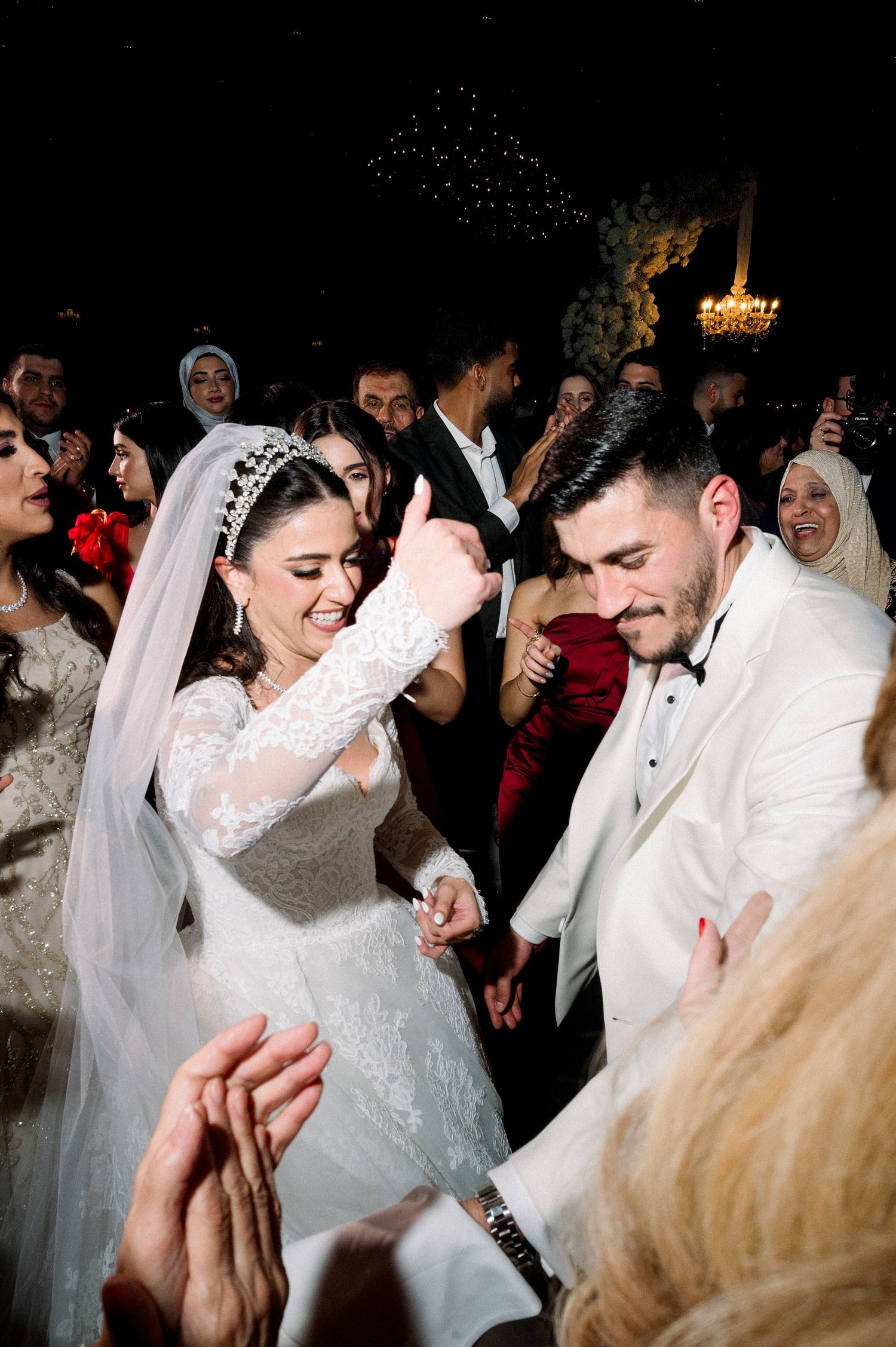 A bride in a white lace gown dances joyfully with guests at a wedding reception celebration.