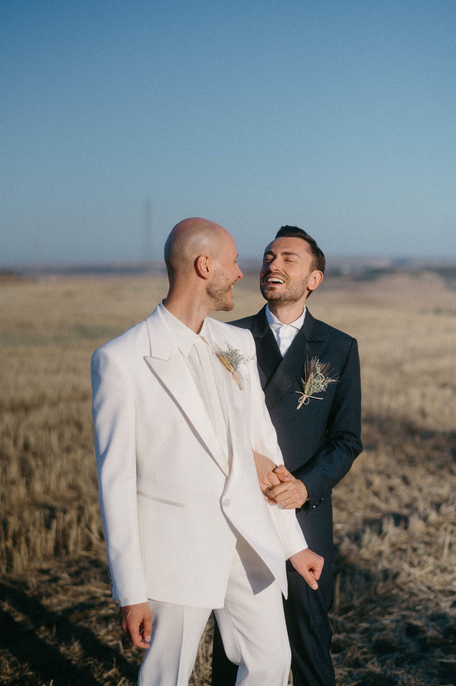 Two people in contrasting white and black suits embrace joyfully in an outdoor field during a wedding photoshoot.