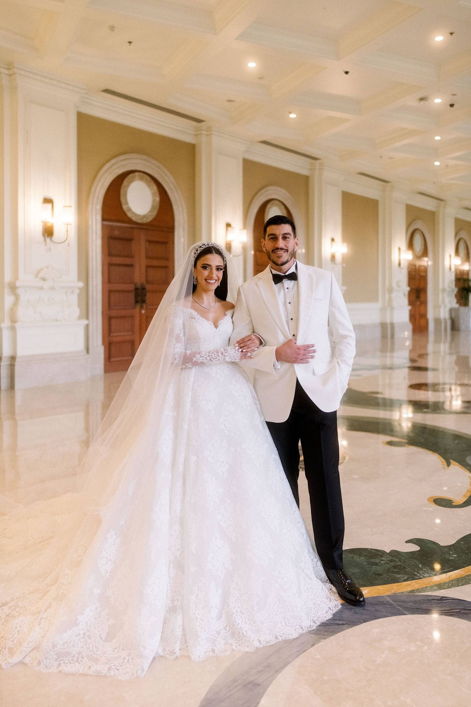 A bride and groom share a romantic moment in an elegant white hallway with ornate columns and warm lighting.