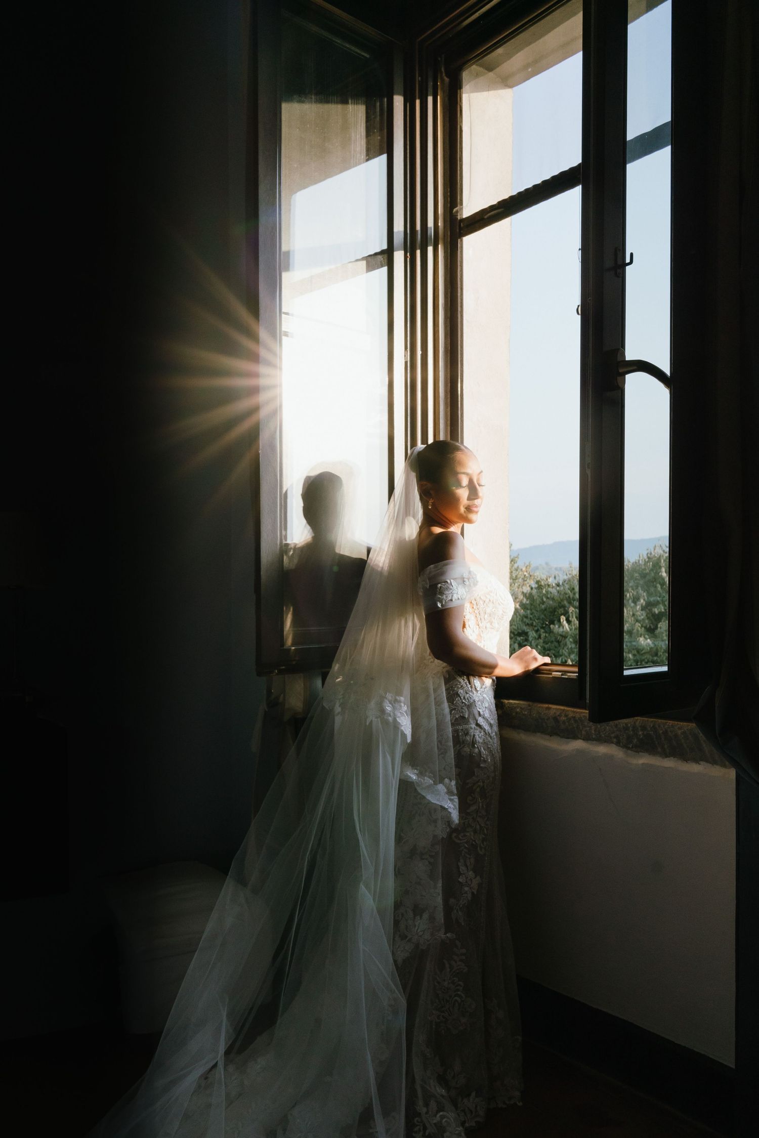 A silhouetted figure in a flowing wedding dress stands by a sunlit window creating a dramatic backlit effect.