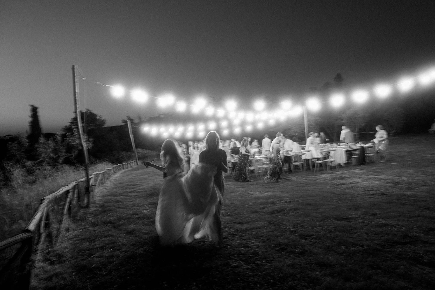 Couple walking toward outdoor wedding reception under string lights at dusk.