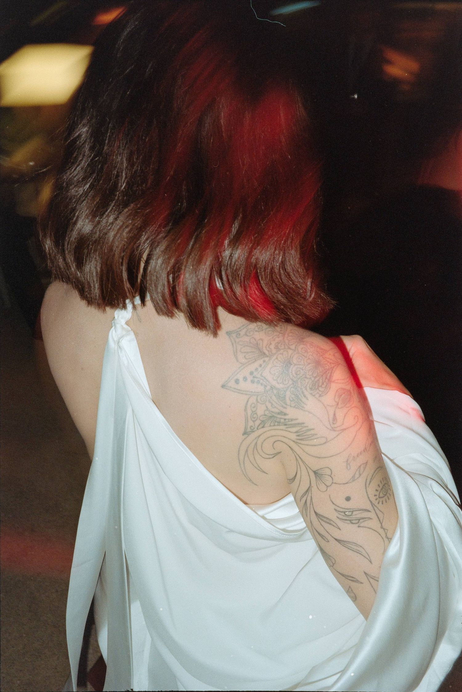 Back view of someone with dark hair and red highlights wearing a white top showing a decorative tattoo on their shoulder.