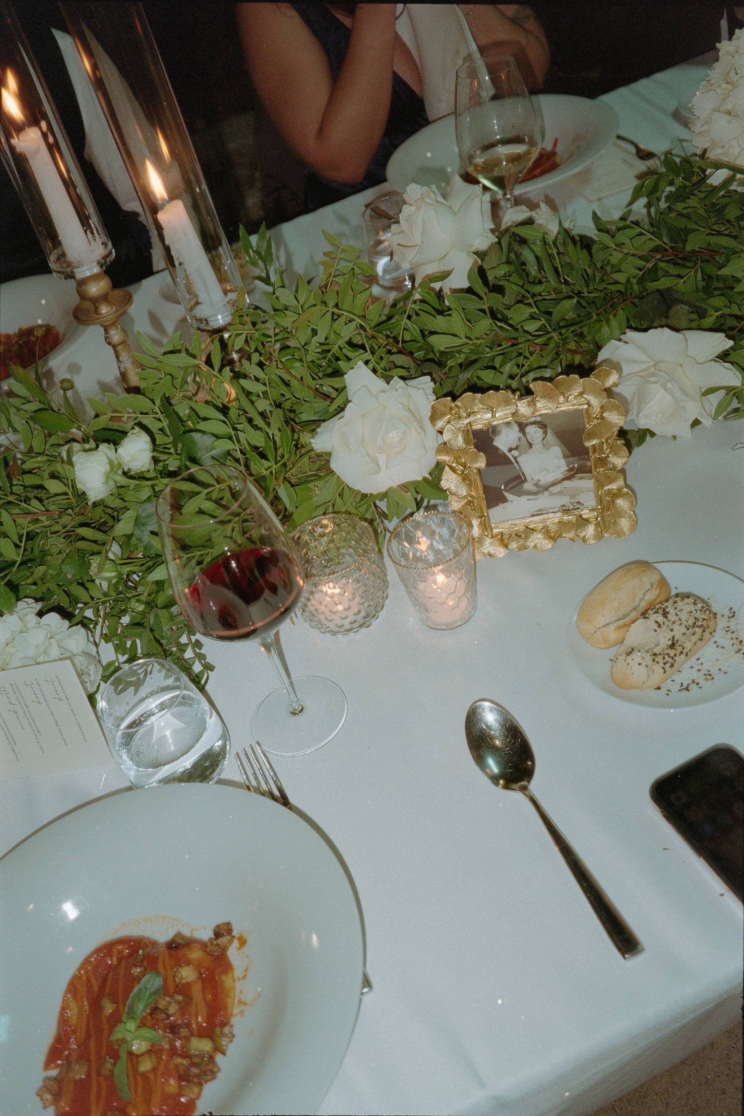 Elegant dinner table setting with candlelight, white roses, greenery, and a pasta dish on white plates.