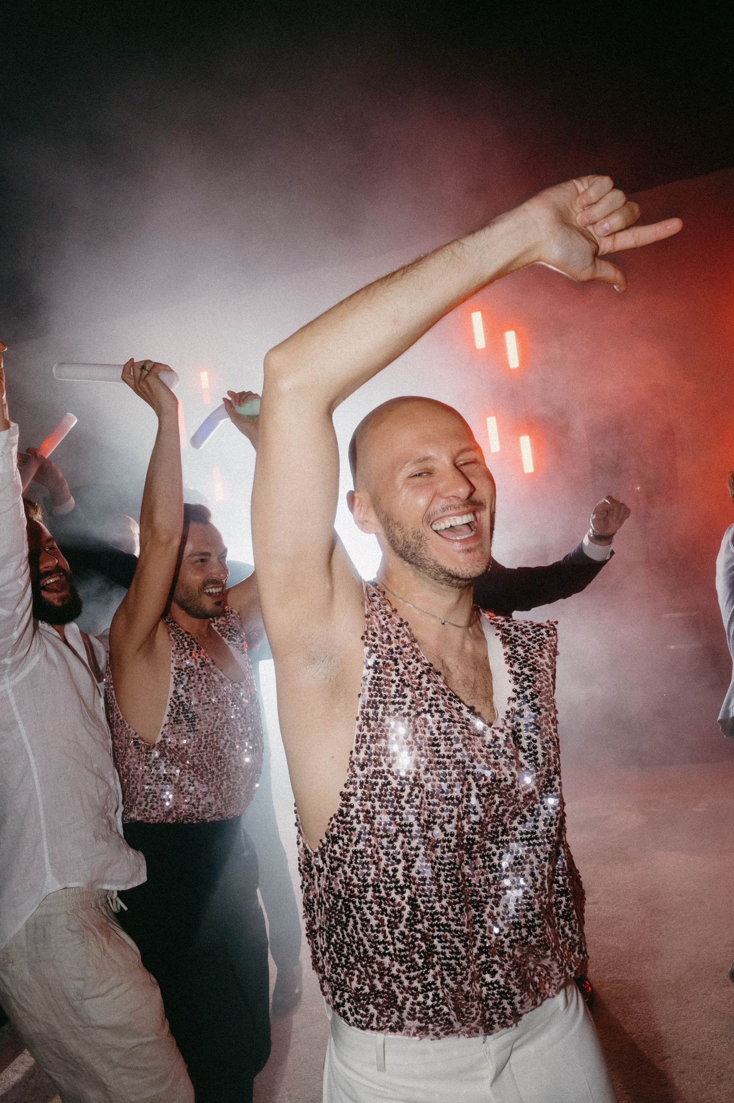 People dancing and celebrating at a party with sparkly outfits in a smoky atmosphere with red lighting.
