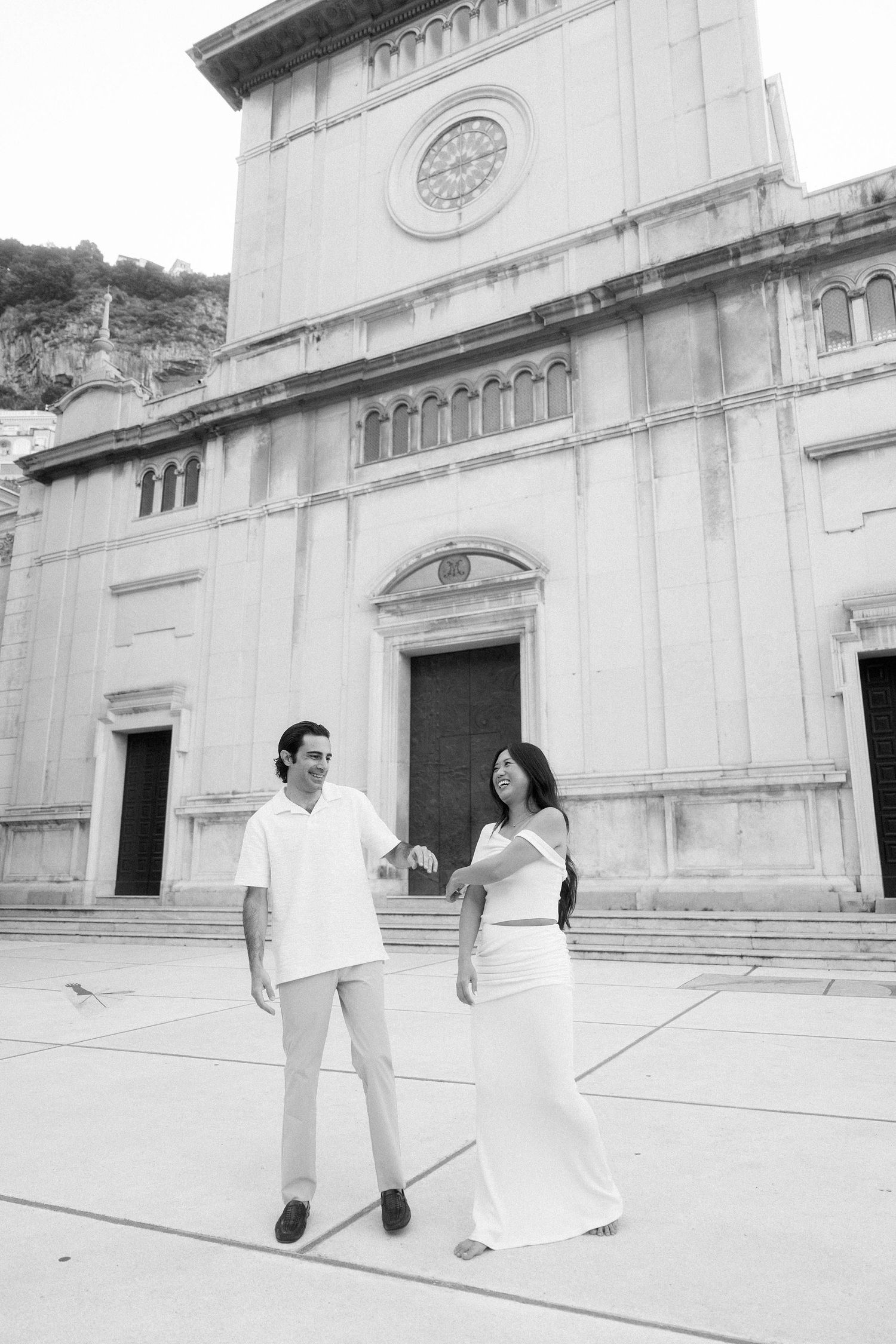 A couple in elegant white attire poses in front of a grand historic church with classical architectural features.