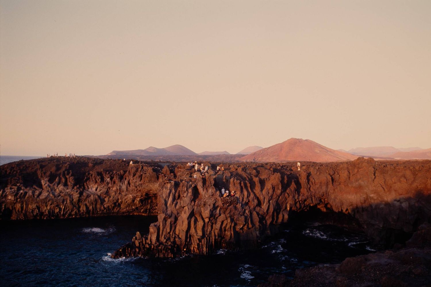 Rugged rocky cliffs and mountains create a dramatic coastline silhouette against a warm sunset sky in a remote landscape.
