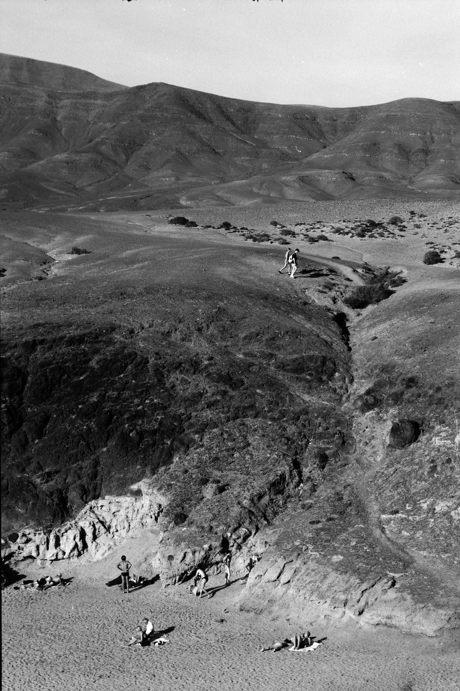 Black and white landscape photograph showing people on a rugged coastal cliff with barren hills in the background.