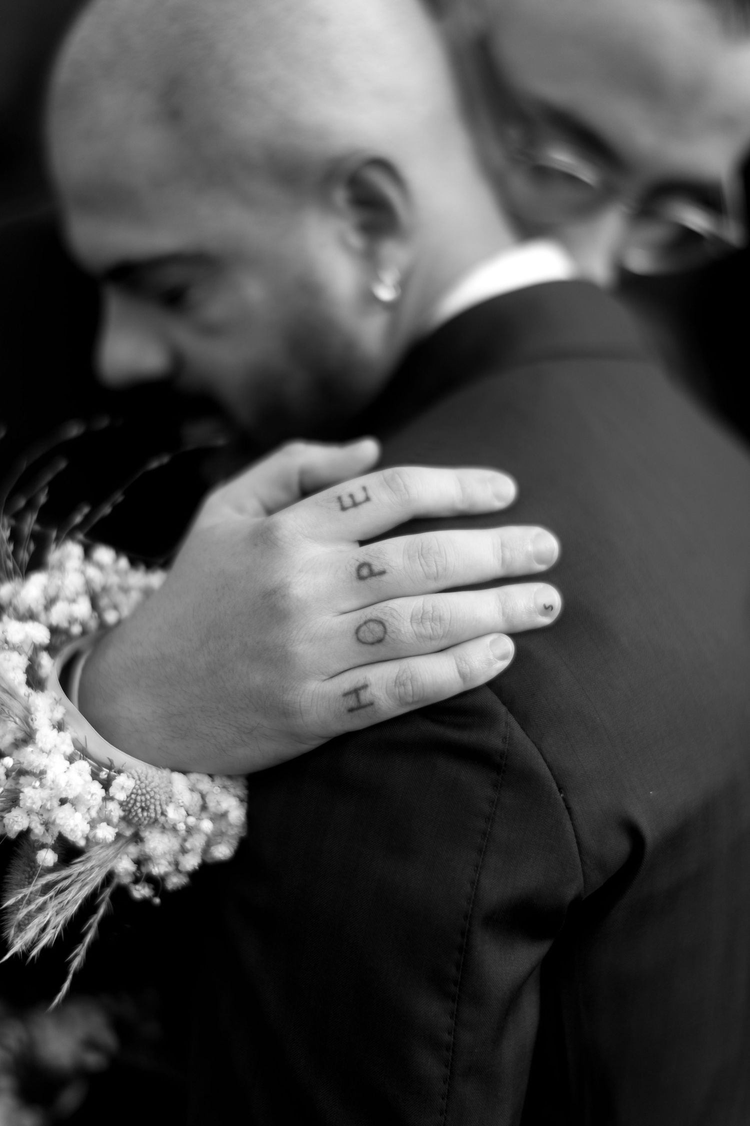 Black and white photo showing a hand with HOPE tattooed on fingers during an embrace at a wedding ceremony.