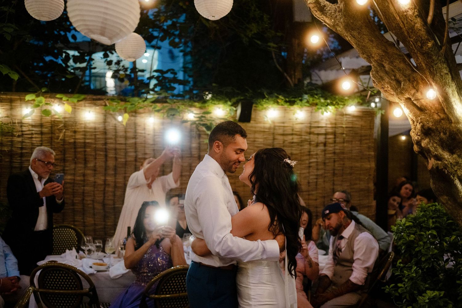 A couple dances together at an outdoor evening reception with string lights and paper lanterns creating a romantic ambiance.