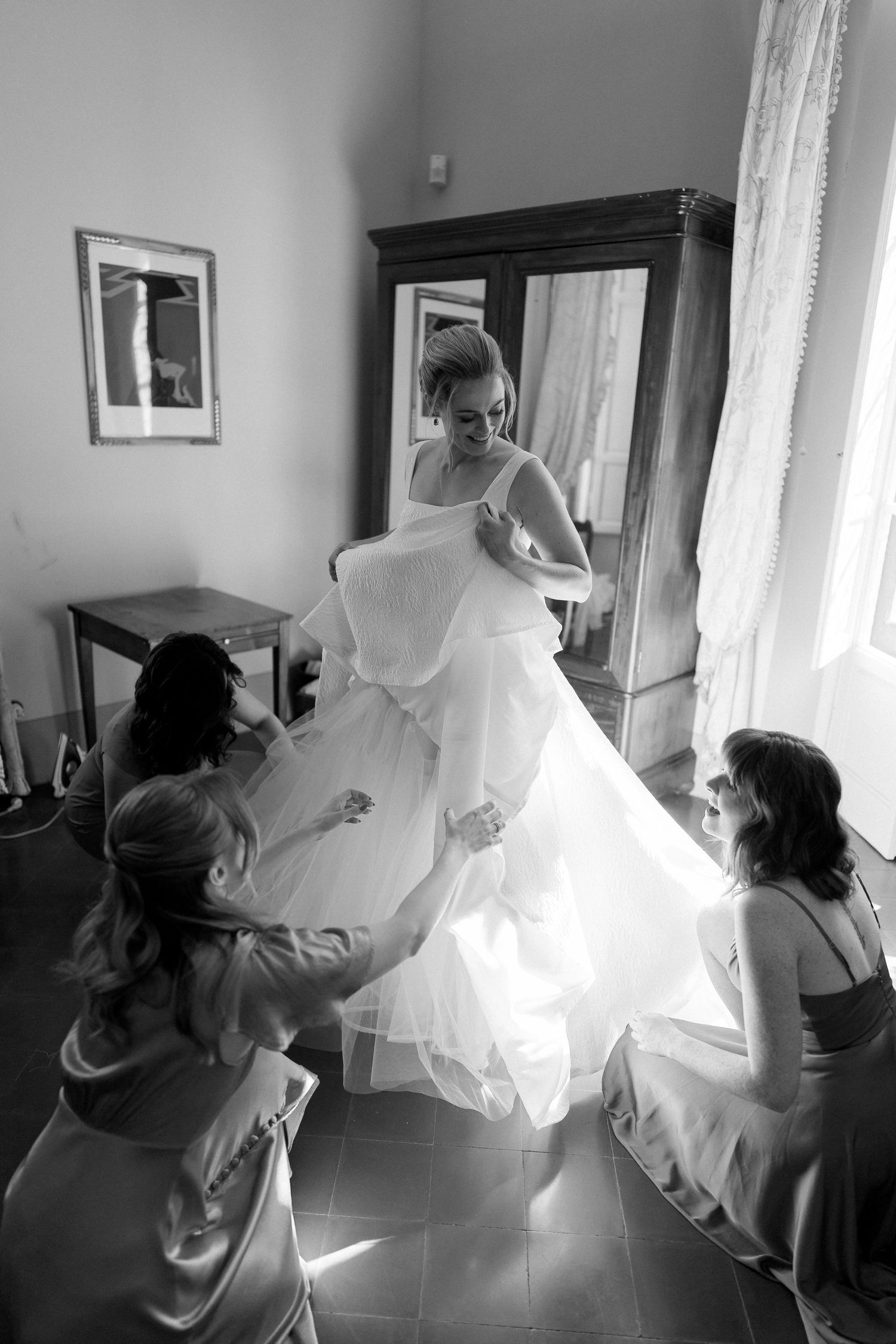 A black and white photograph captures the intimate moment of bridesmaids helping adjust a flowing wedding gown in sunlit room.