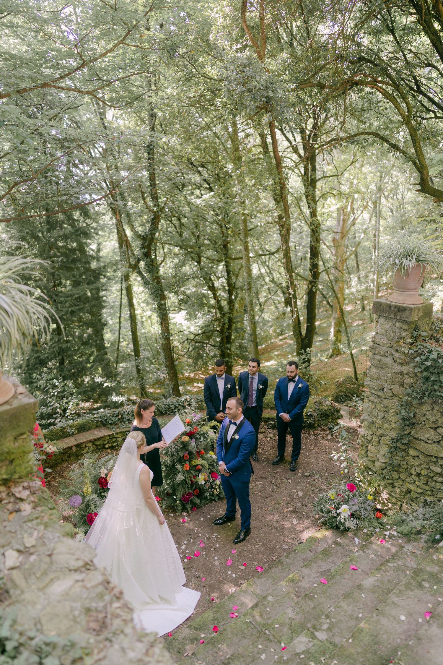 A romantic outdoor forest wedding ceremony with pink rose petals scattered along a wooded path.