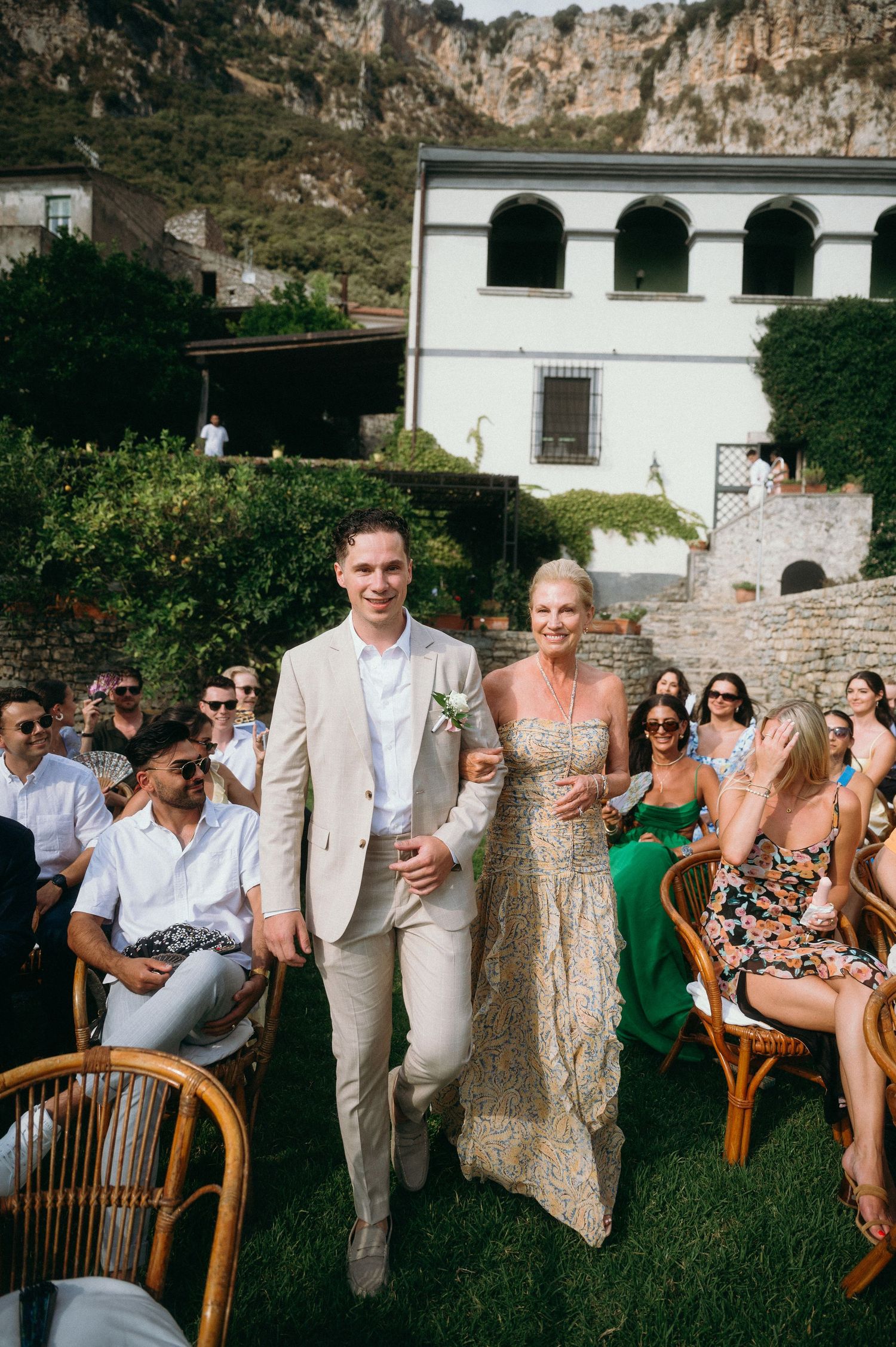 Wedding guests in light summer attire gather outdoors against a scenic stone wall backdrop for group photos.