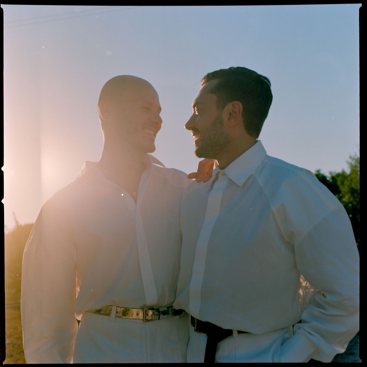 Two figures in white shirts silhouetted against a bright sunset sky in a romantic outdoor setting.