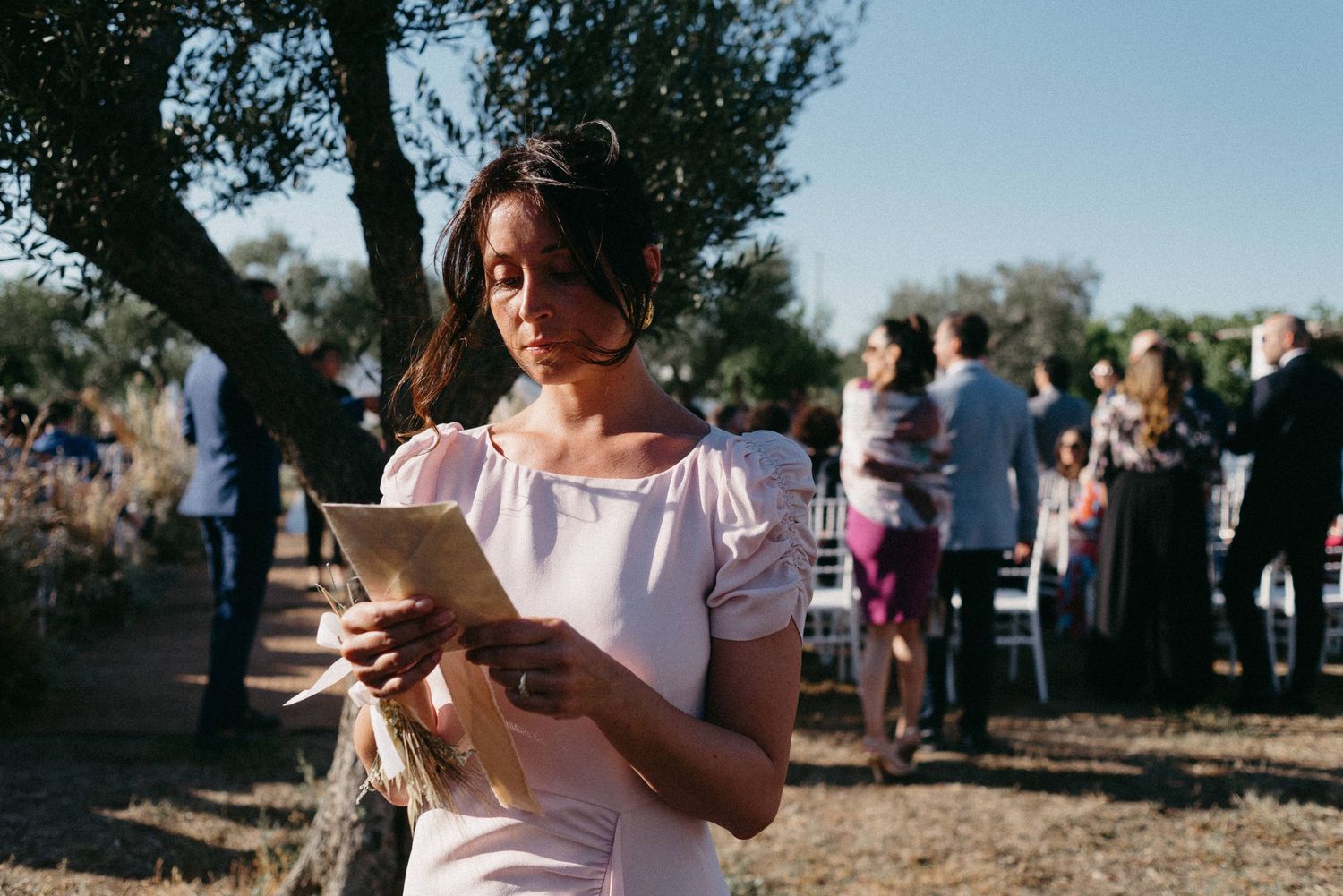 A person holds a smartphone while standing outdoors in a sunny park setting with trees and people in the background.