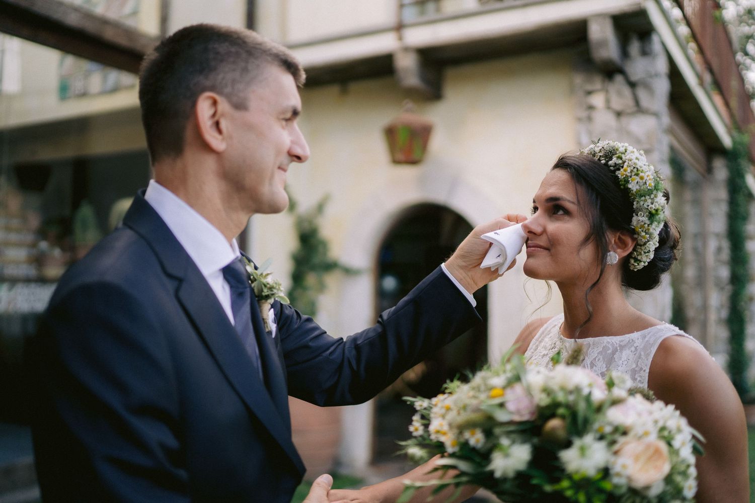 A tender wedding moment as a groom gently wipes away tears from an emotional bride holding a pastel bouquet.
