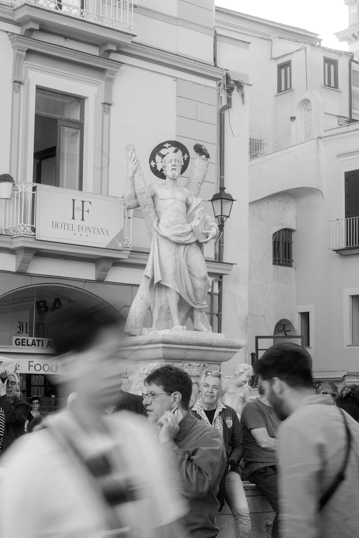 Black and white photo of a classical statue in an Italian plaza with blurred tourists walking past in the foreground.