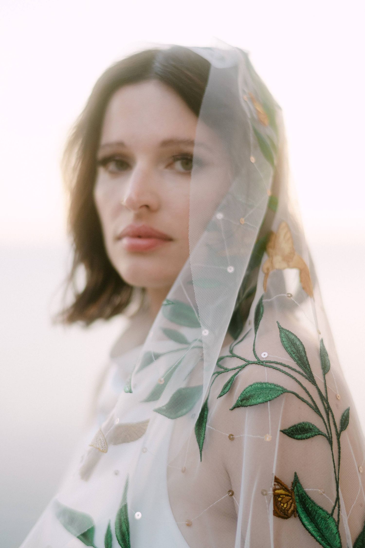Ethereal portrait through sheer floral veil with embroidered green leaves and butterfly details in soft lighting.