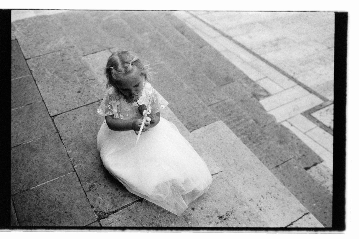 A young child in a white flowing dress lies on concrete pavement while looking up at the sky, captured in black and white.