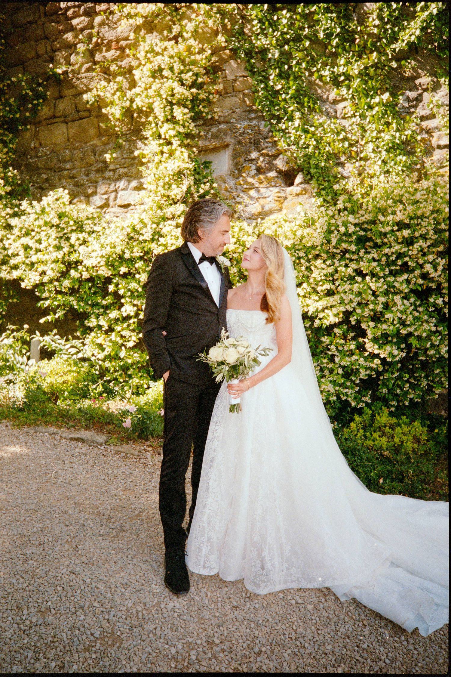 Bride and groom share a romantic moment in a lush garden, she in a strapless lace gown holding a white bouquet.