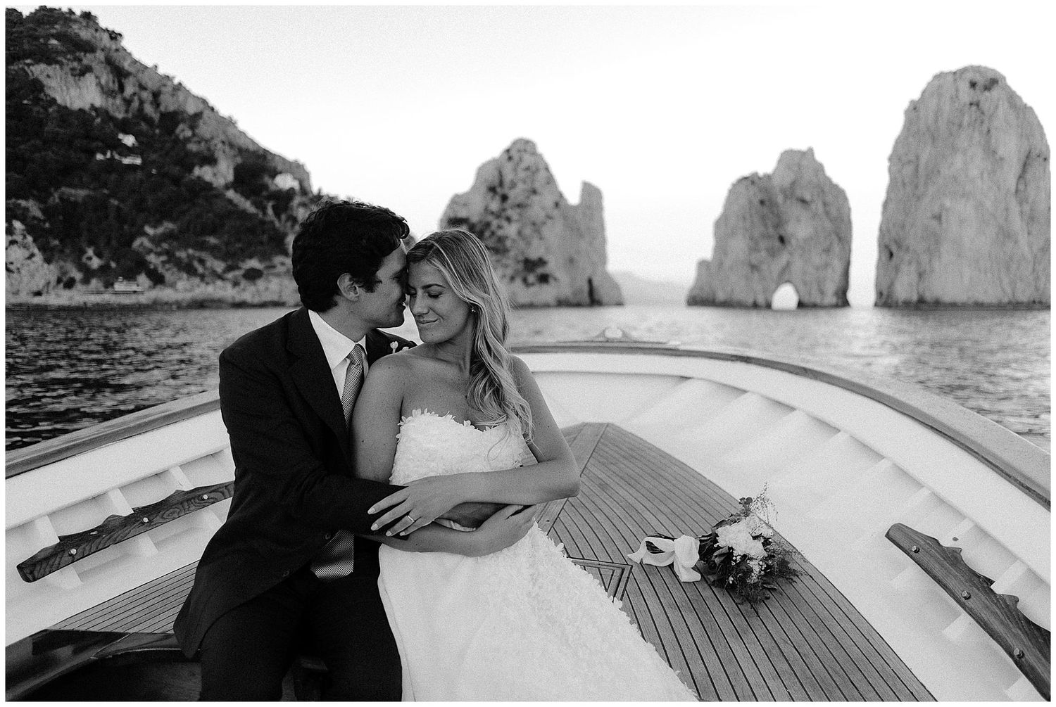 Black and white photo of bride and groom sharing romantic moment on boat deck with Capri's Faraglioni rocks visible.