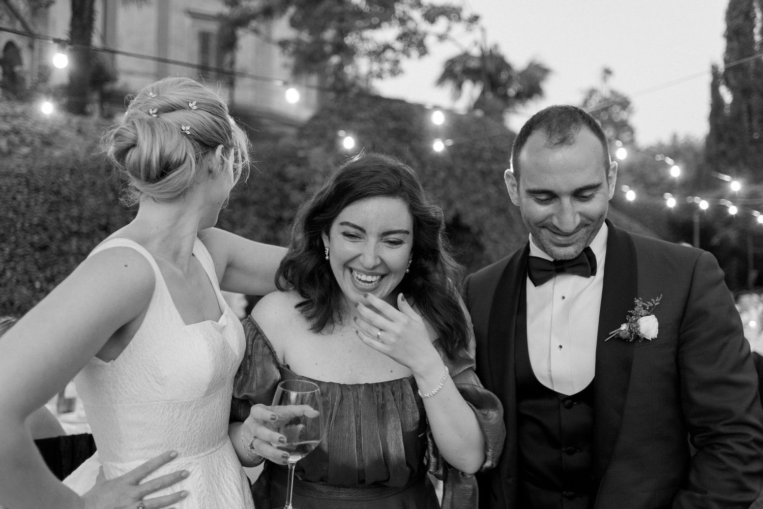 Three people share a candid moment at an evening event, laughing together while holding drinks.