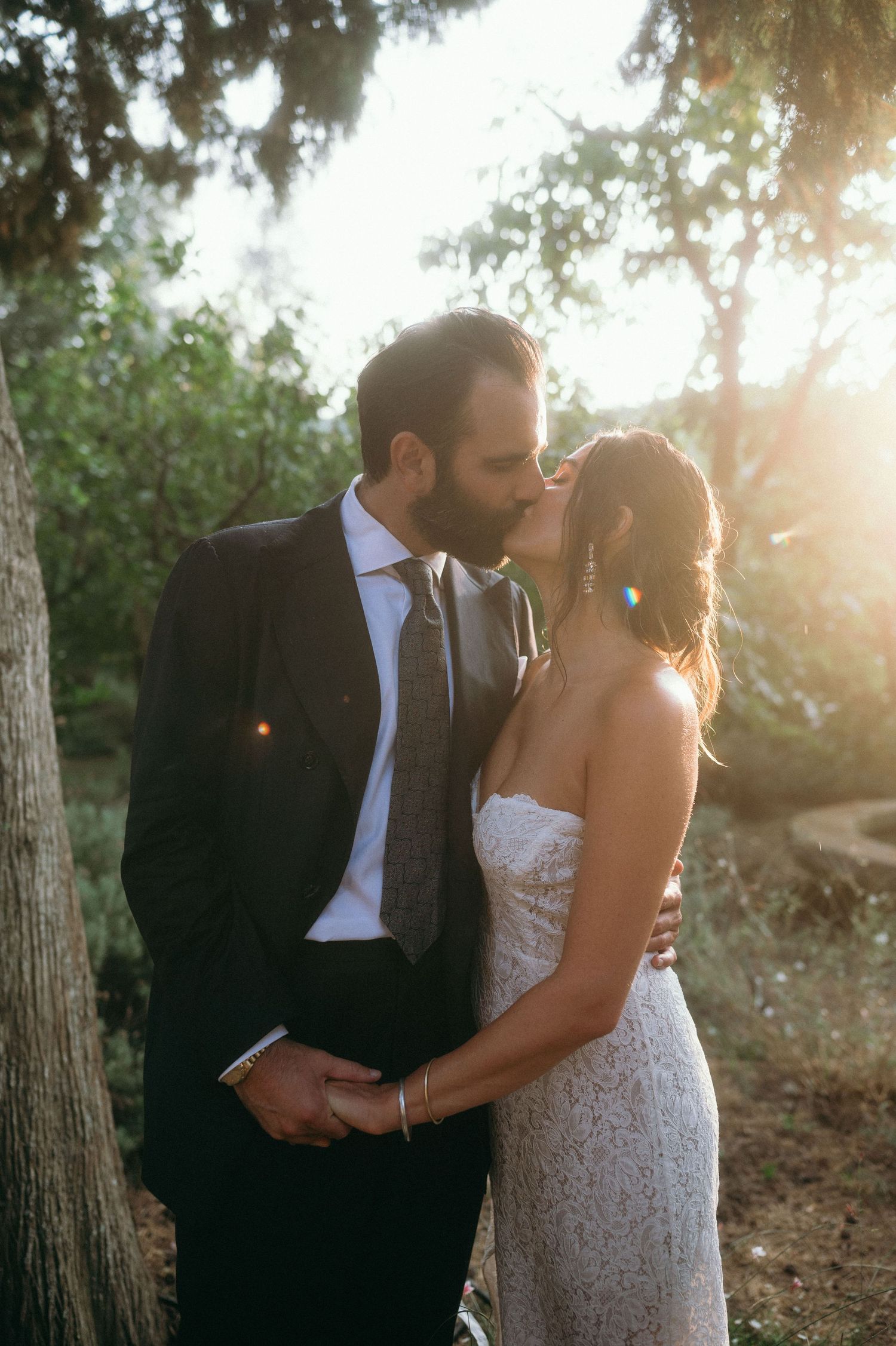 Newlywed couple sharing an intimate kiss in a sun-dappled forest setting, with golden light filtering through trees.