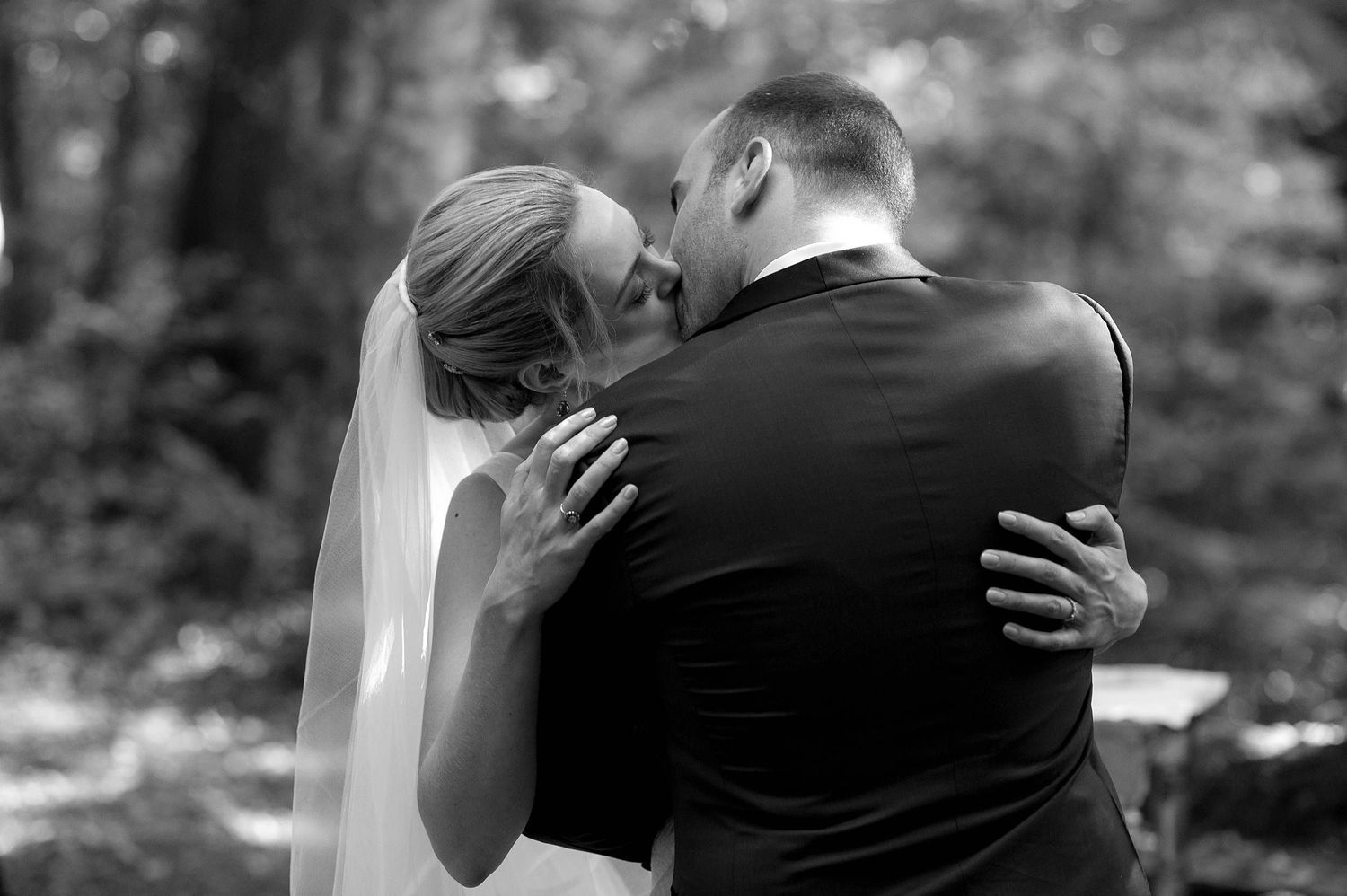 A romantic black and white wedding photo showing a couple embracing in a wooded outdoor setting.