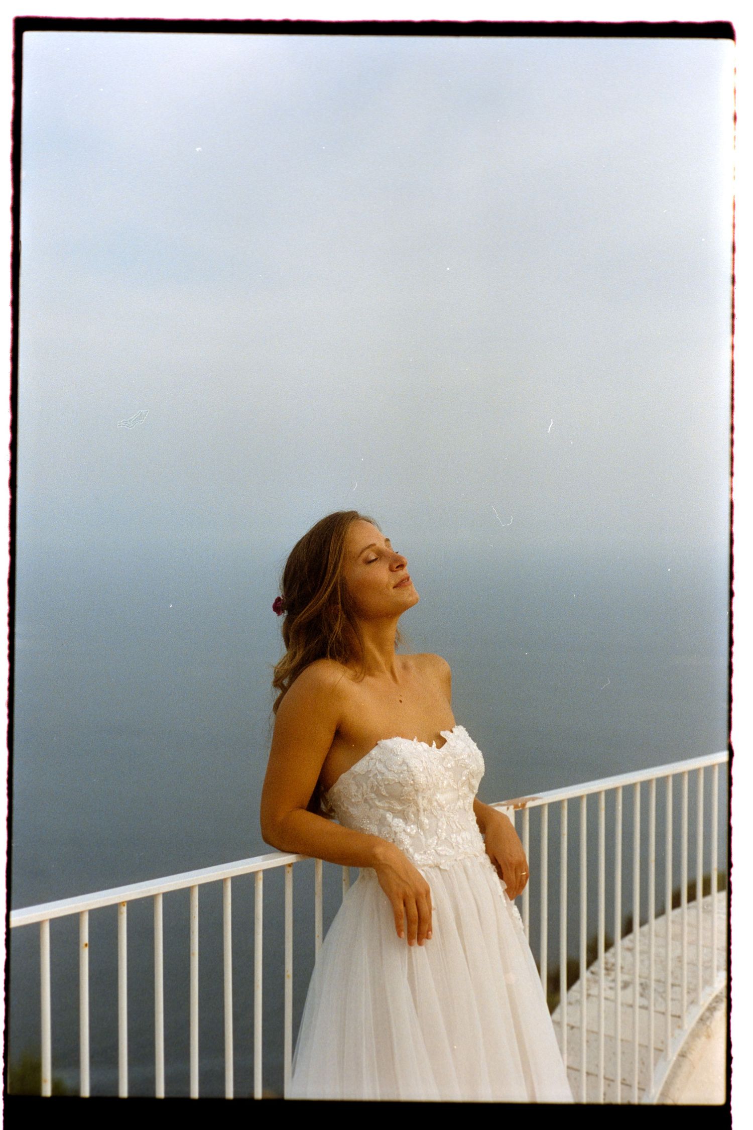 A person in a white strapless wedding dress poses on a balcony overlooking a misty view.