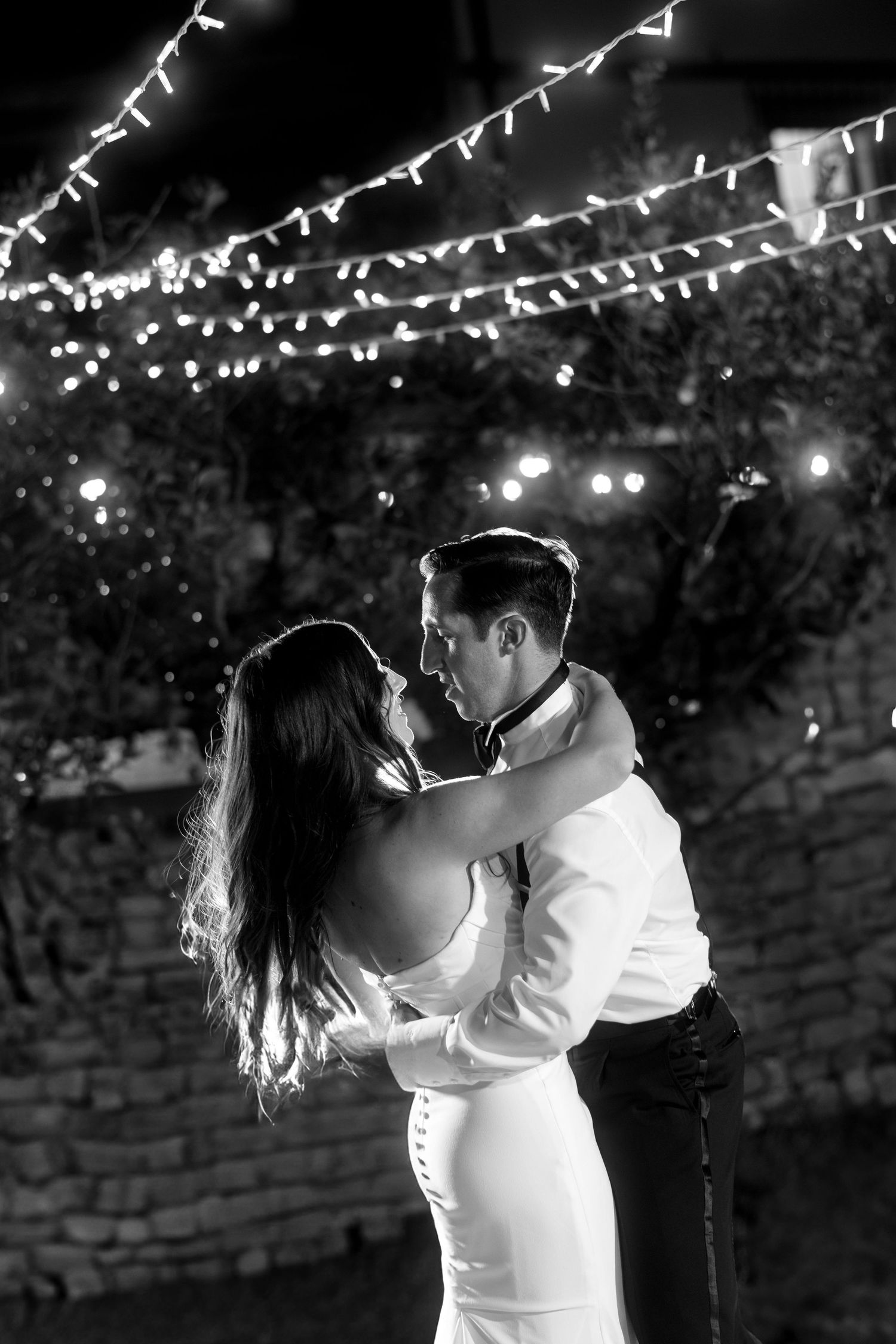A romantic couple shares their first dance under twinkling string lights at an outdoor evening wedding reception.