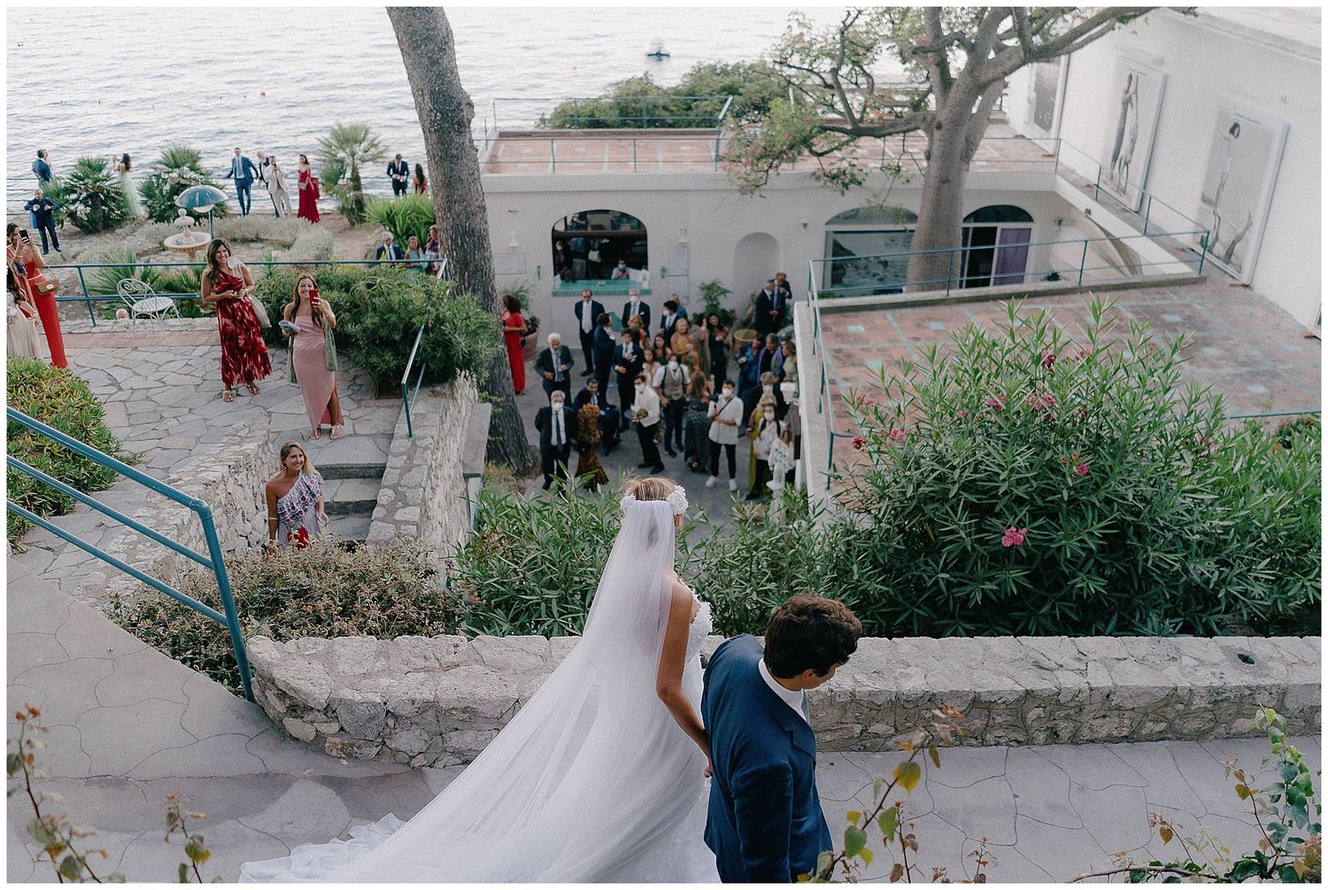 A bird's eye view of wedding guests gathering outside while newlyweds descend garden stairs.