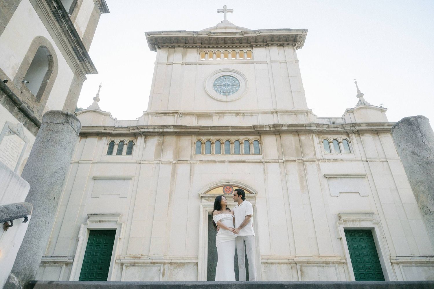 A couple embraces in front of a white Catholic church facade with green doors and a cross on top.
