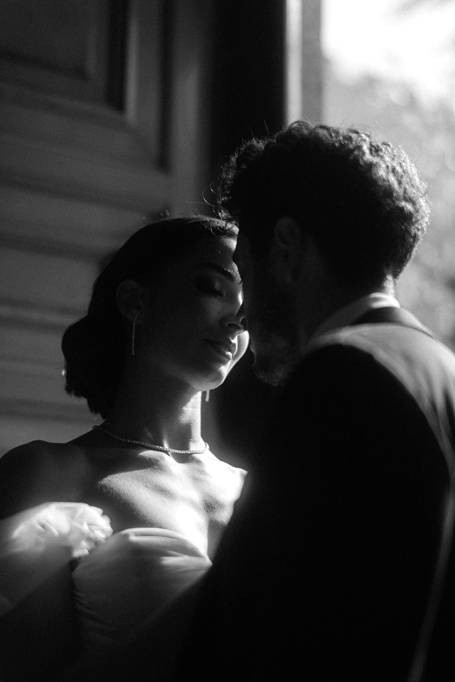 Black and white intimate wedding portrait of couple in romantic lighting by window.