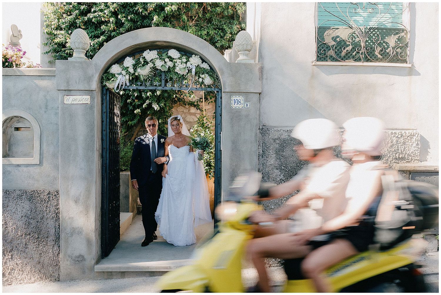 Couple stands in a stone archway decorated with white flowers as cyclists blur past in motion on the street.